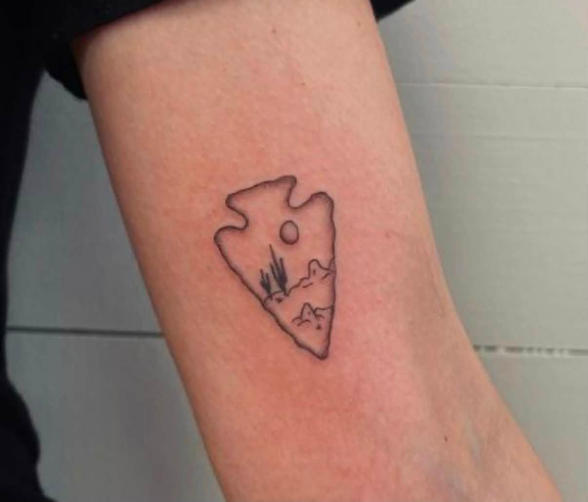 Close-up of a forearm with a small black outline tattoo of a landscape and sun.