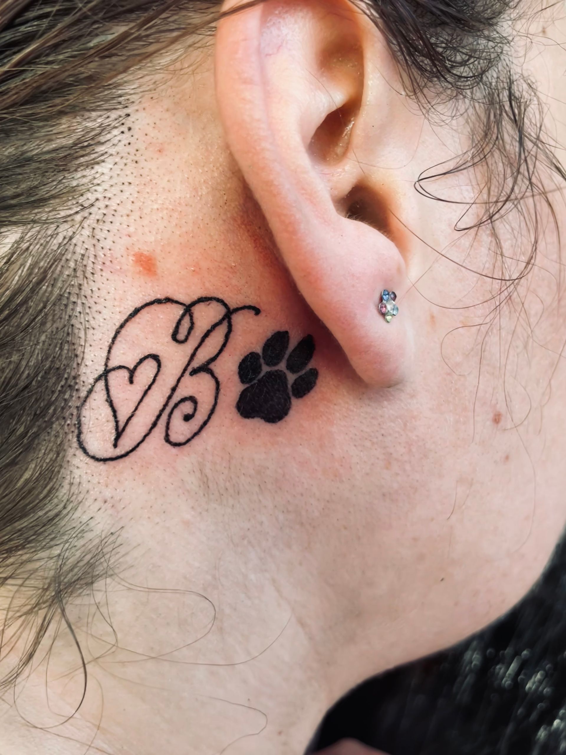 Close-up of an ear with a heart monogram tattoo and a black paw print tattoo behind it