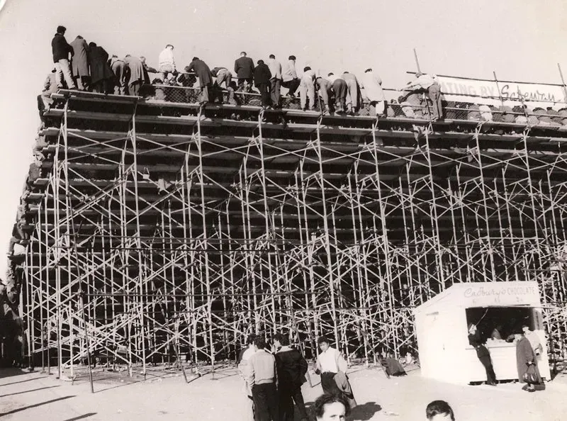 A historical image in black & white of a grandstand scaffold built with people 