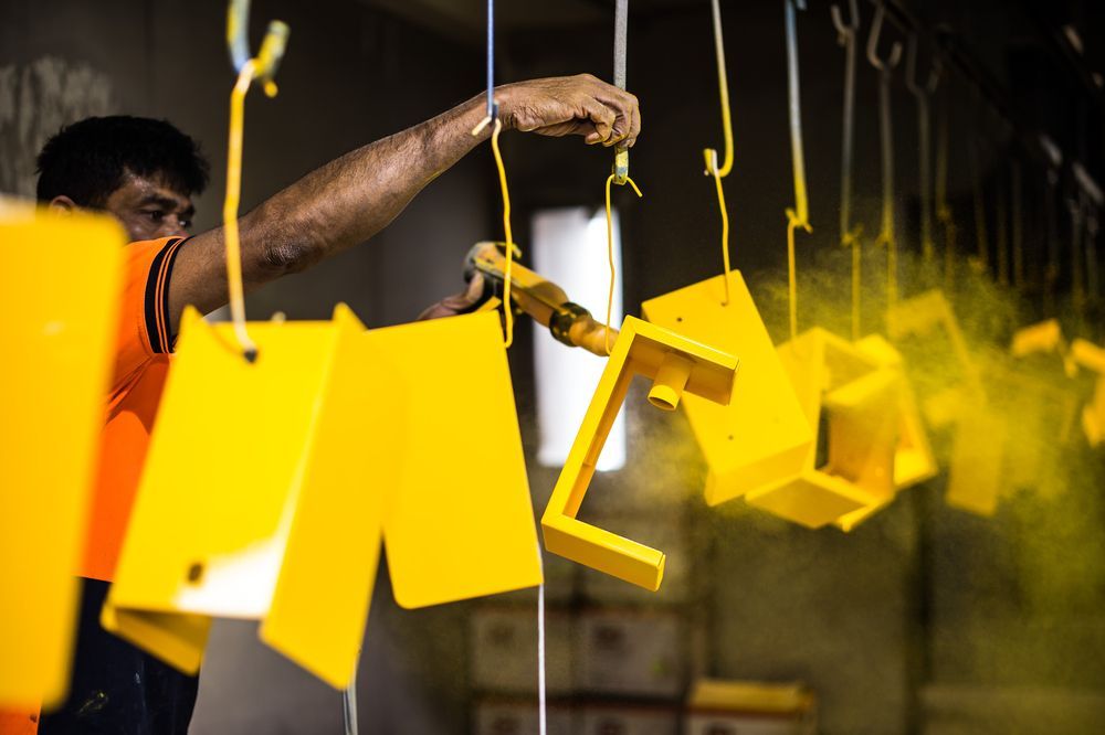 A Man Is Painting Yellow Metal Pieces in A Factory — Alamo Powder Coaters in Unanderra, NSW