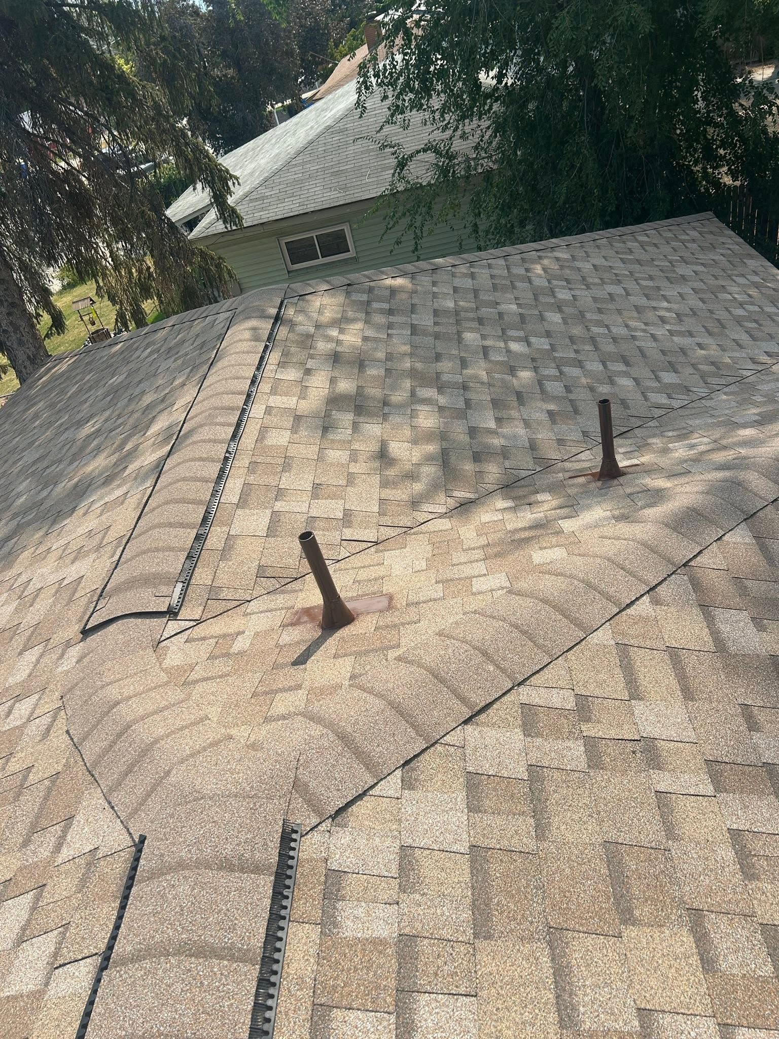 Rooftop covered in gray shingles with two vent pipes, viewed through trees.