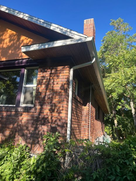 Brick house exterior with a chimney, sloped roof, and leafy trees in bright sunlight