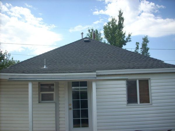 Back view of a beige house with white trim and a dark shingled roof under a partly cloudy sky