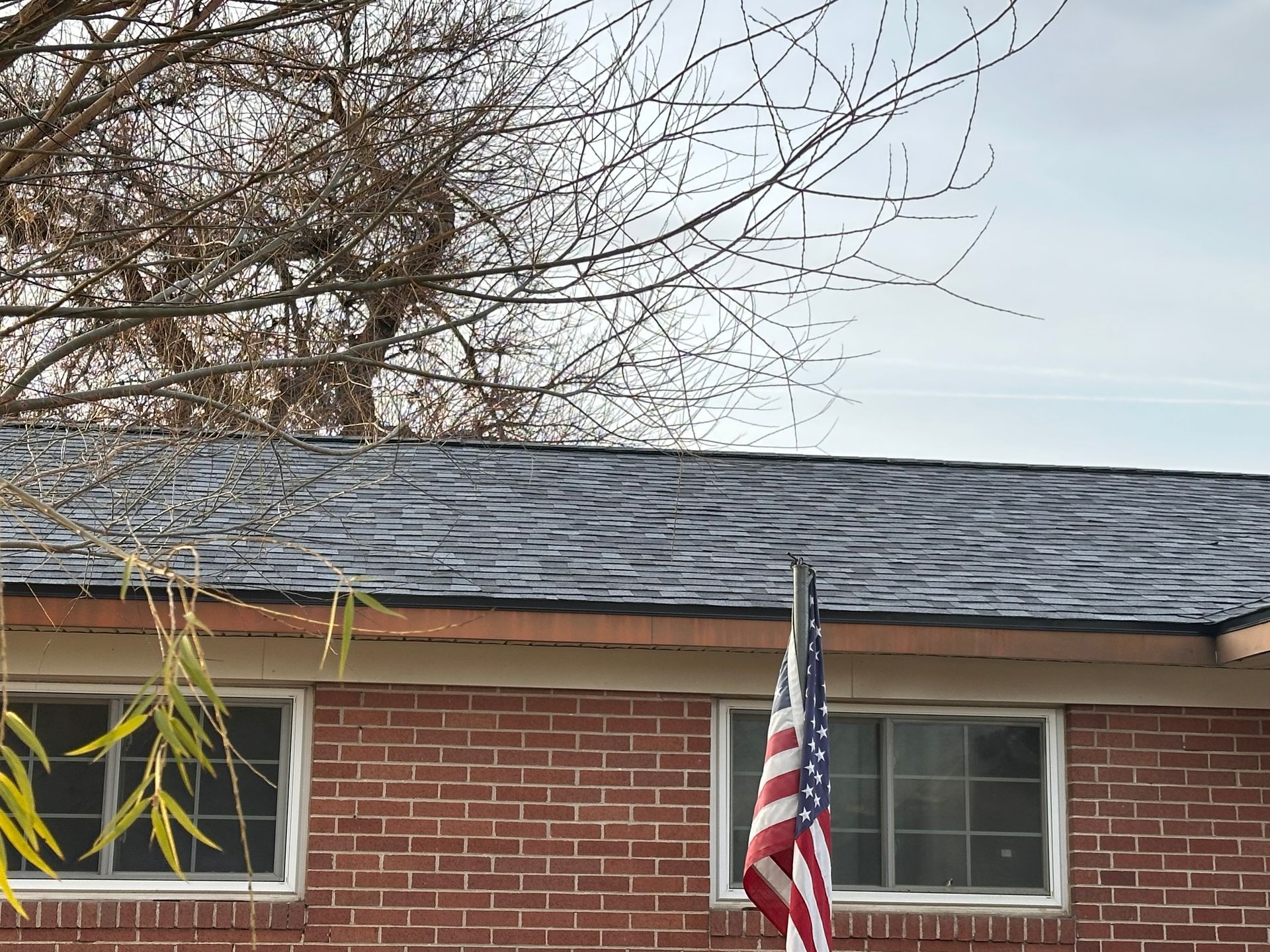 Brick house roof with American flag hanging by a window and bare tree branches overhead