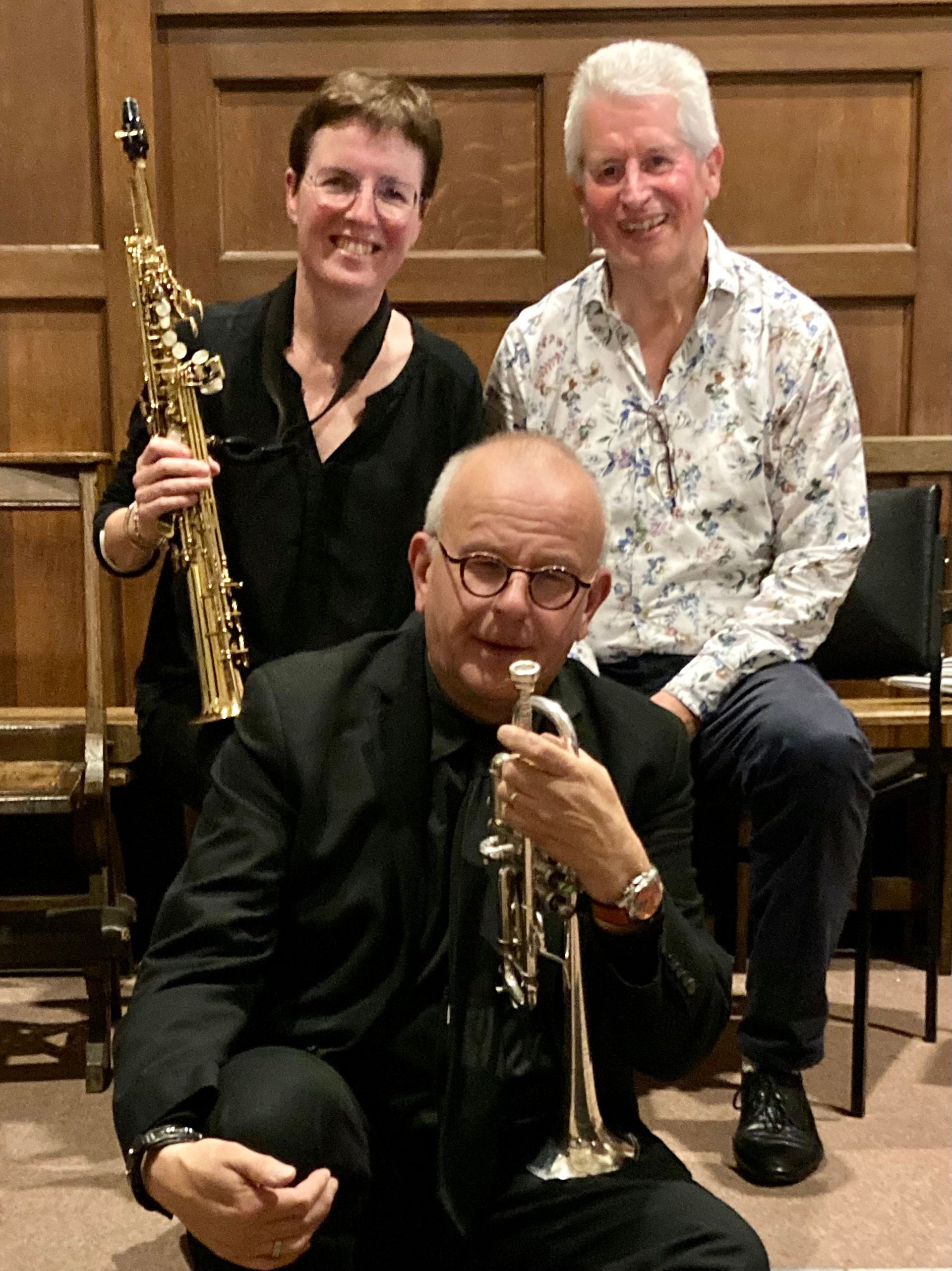 Drie muzikanten: een trompettist op de voorgrond, een saxofonist en een lachende man op de achtergrond. Binnenlocatie.