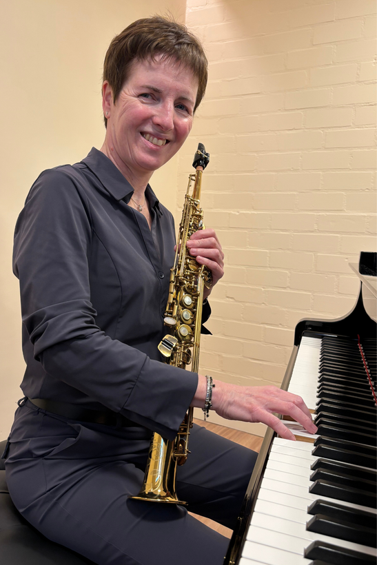 Vrouw zit aan een piano en houdt een saxofoon vast. Ze glimlacht, binnen.
