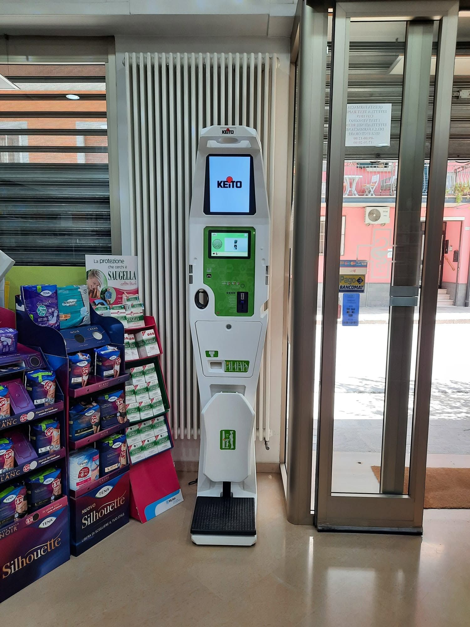Chiosco self-service all'ingresso di un negozio, accanto a uno scaffale di snack e a una porta a vetri aperta.