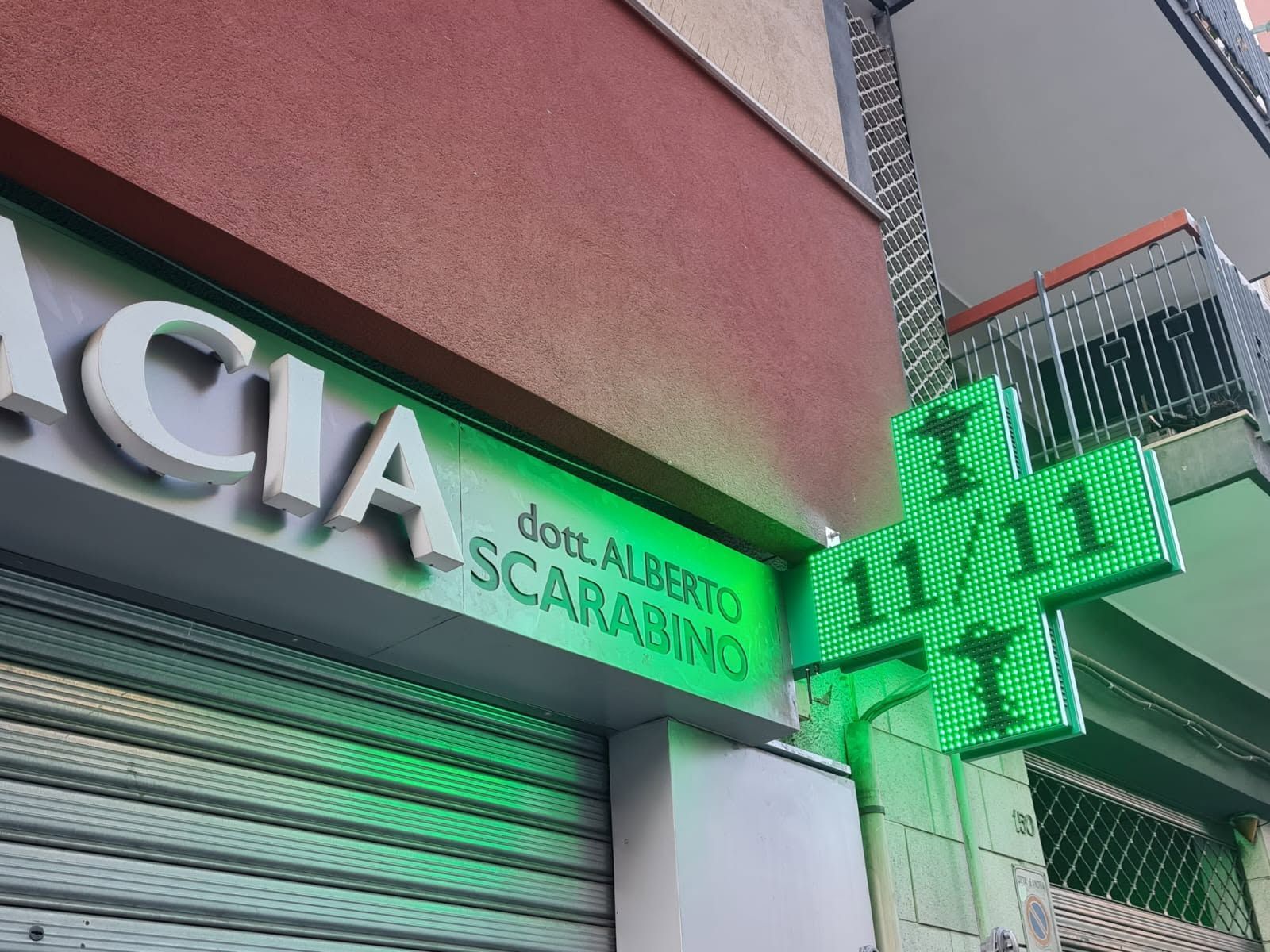 Insegna di farmacia su strada con croce verde illuminata e tendalino sotto la sporgenza di un edificio.