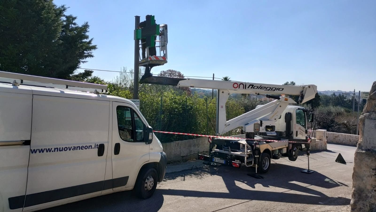 Un furgone bianco e un camion con piattaforma elevatrice sono parcheggiati all'aperto su una strada soleggiata vicino a una siepe.