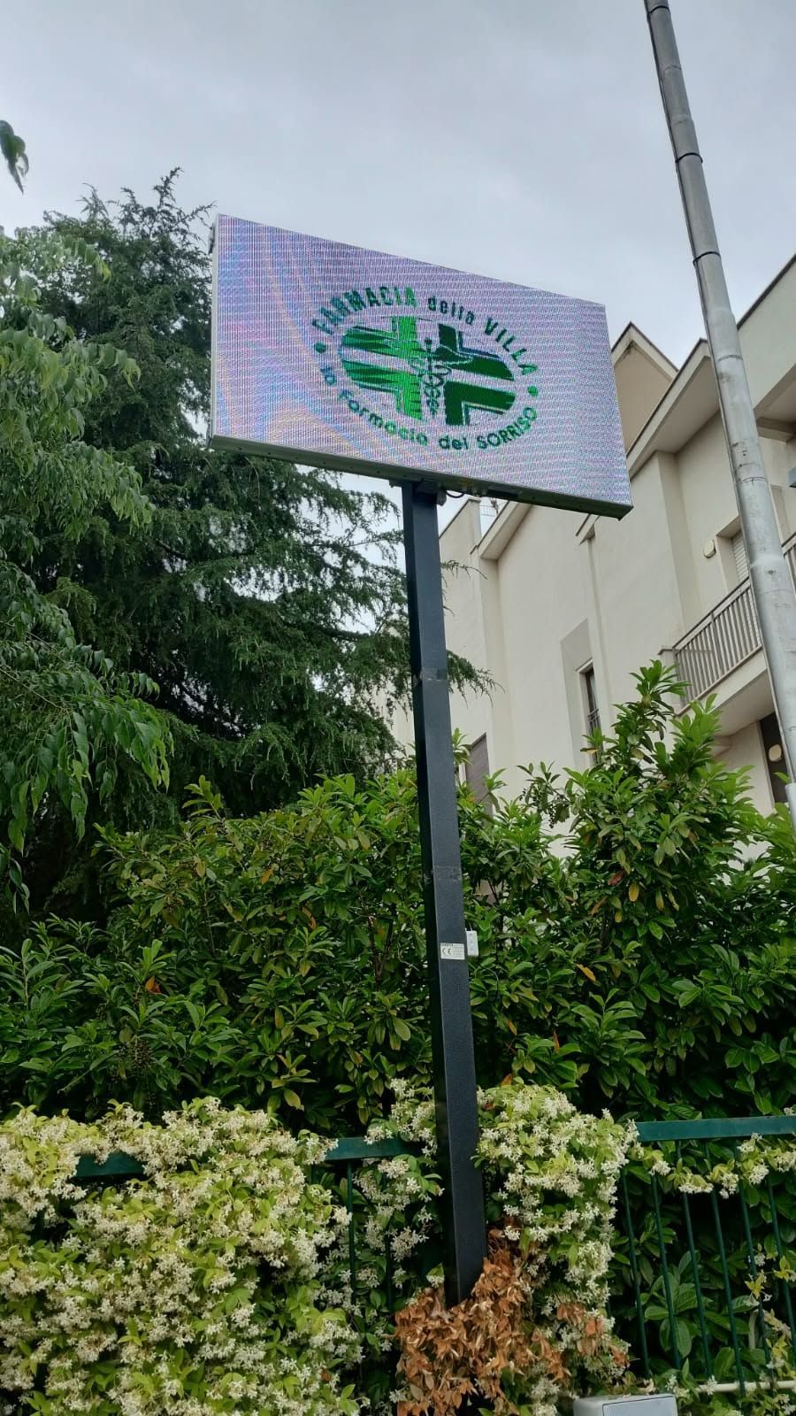 Cartello verde e bianco su un palo sopra dei cespugli, con un logo circolare a forma di croce all'esterno.