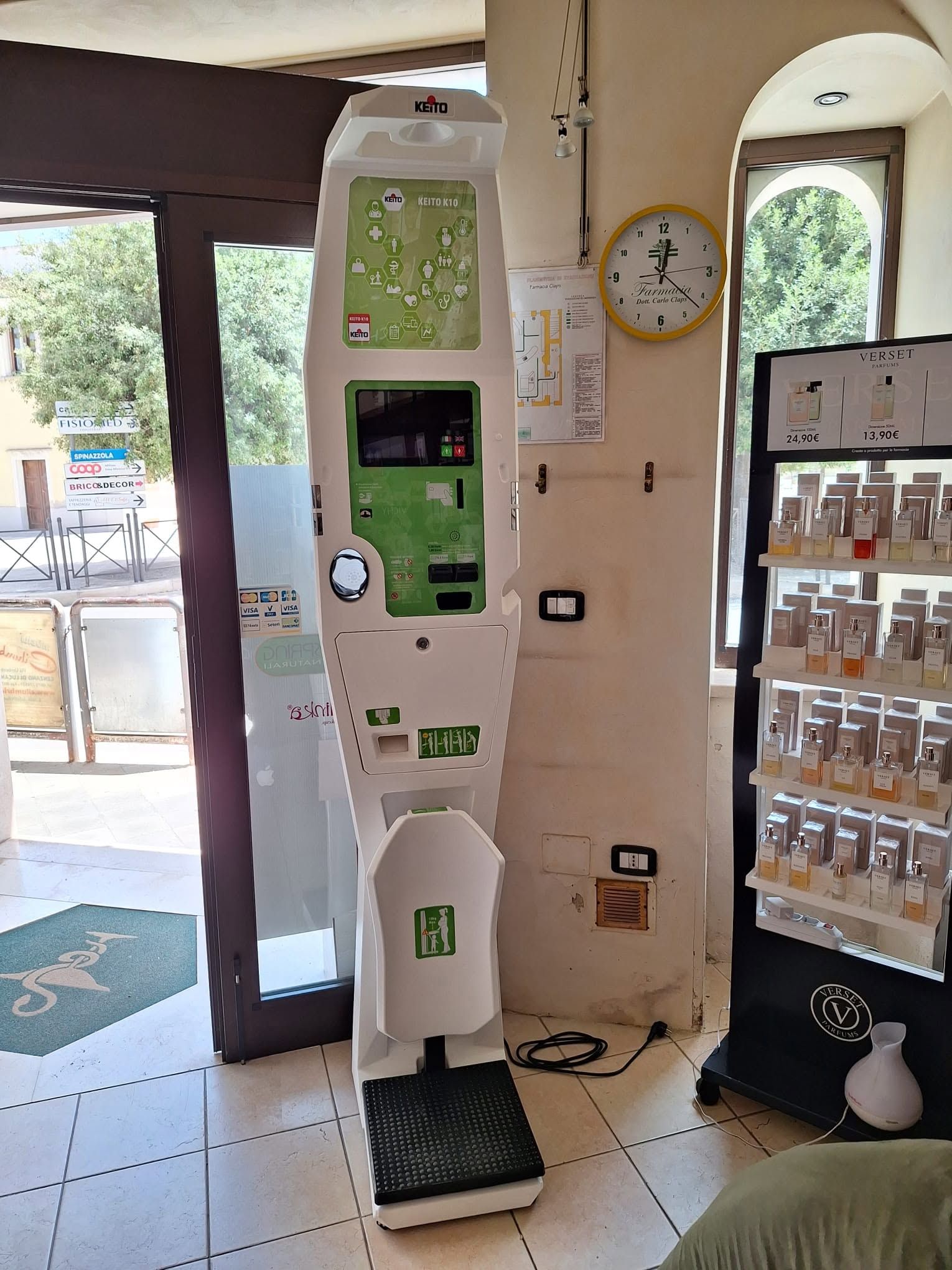 Chiosco interno accanto a un distributore automatico in una hall piastrellata, con un orologio a muro e una porta aperta verso l'esterno.