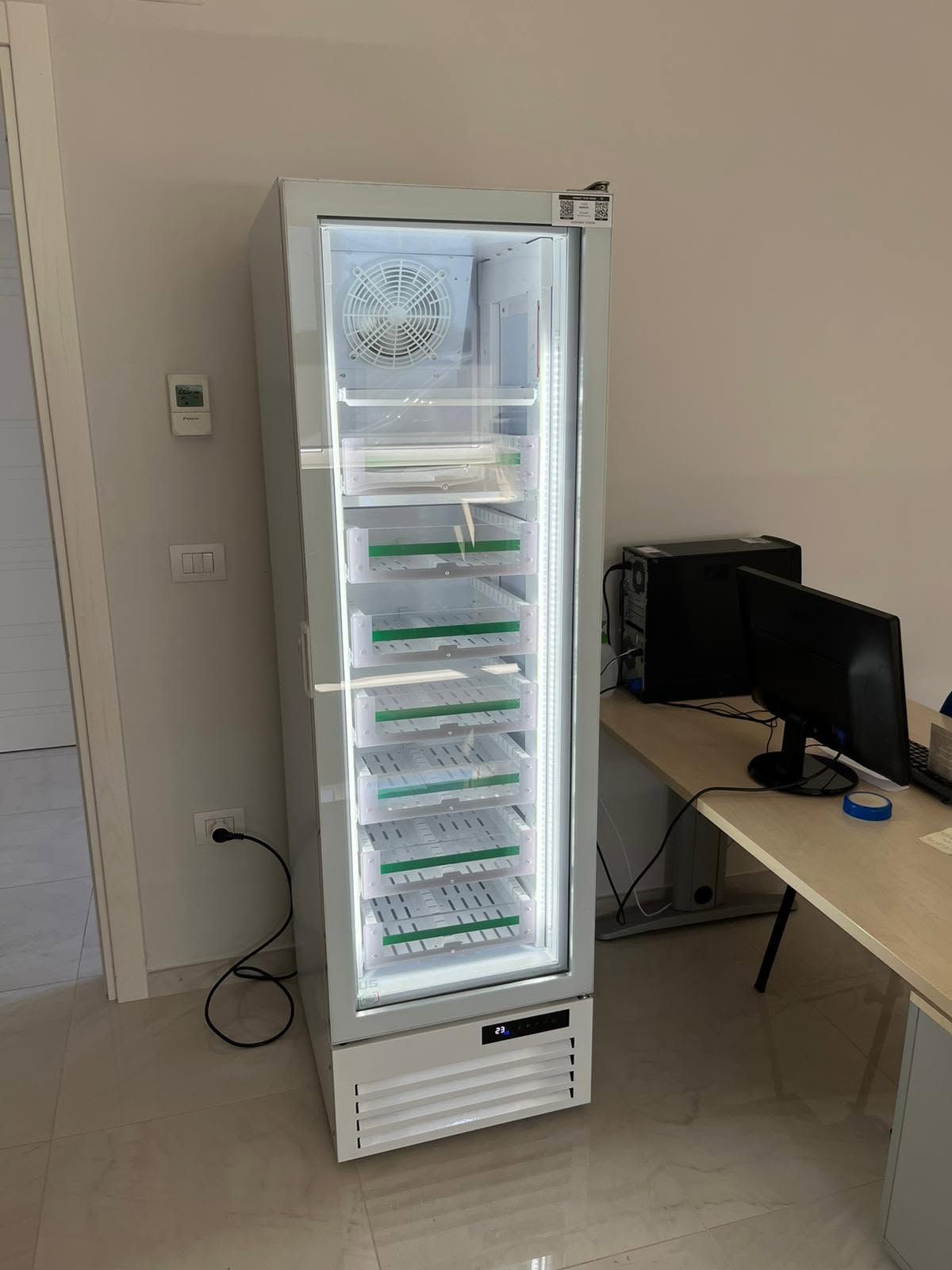 Frigorifero o congelatore alto e bianco, posizionato in ufficio accanto a una scrivania con monitor.