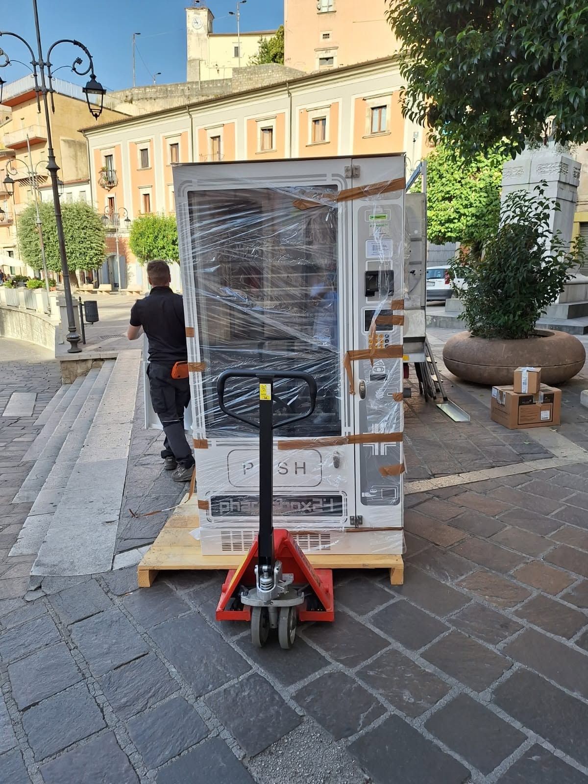 Un elettrodomestico imballato è posizionato su un transpallet in una piazza lastricata in pietra, con una persona che cammina nelle vicinanze.
