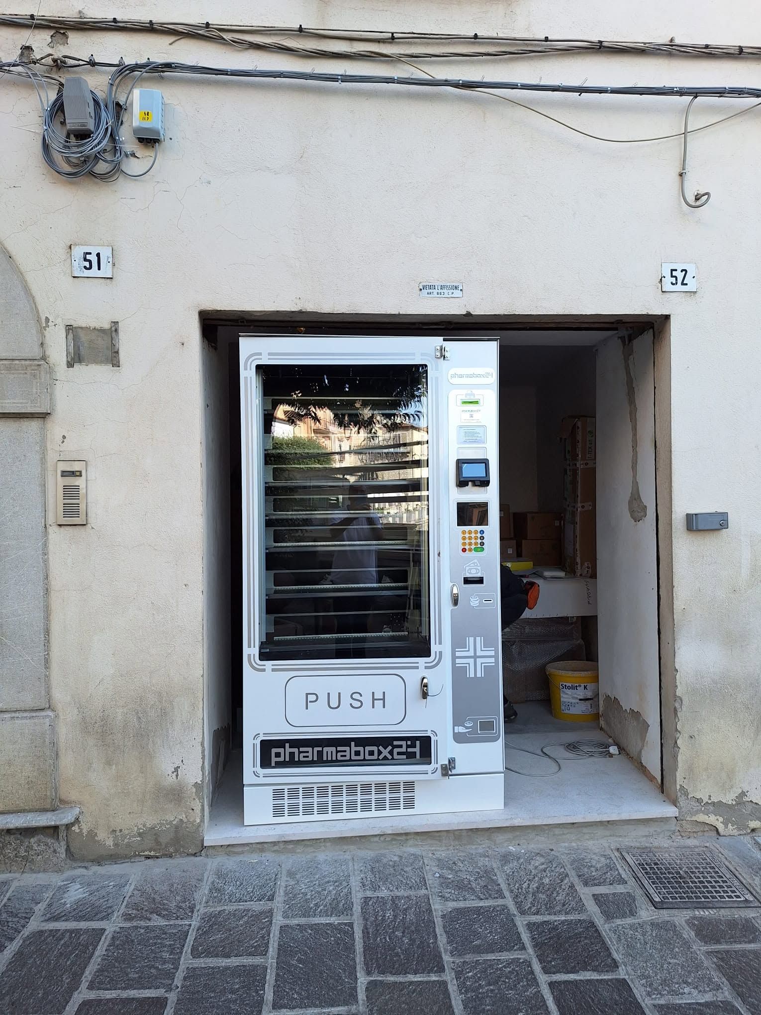 Piccola vetrina con un distributore automatico bianco incastonato in un muro di pietra, su una strada asfaltata.