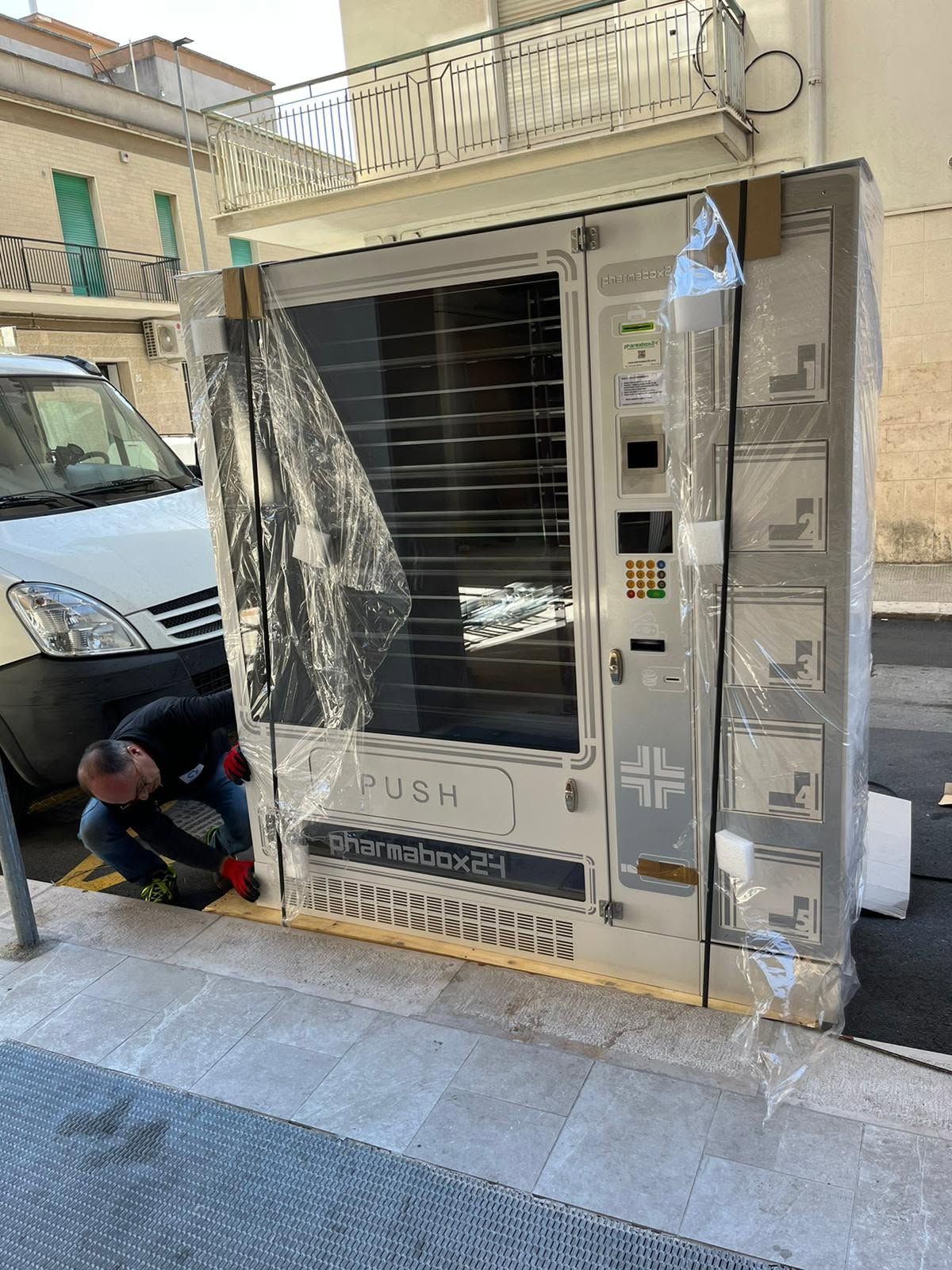 Un distributore automatico viene riparato in strada, avvolto nella plastica, con un operaio inginocchiato accanto.