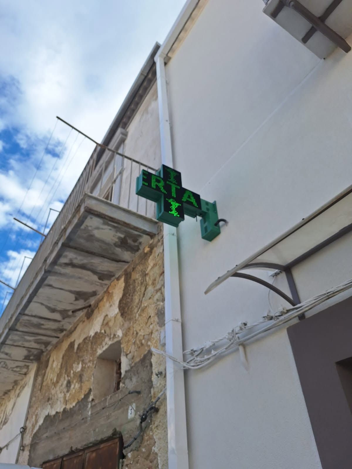 Una croce illuminata di verde, montata sulla parete bianca di un edificio, si staglia contro un cielo azzurro.