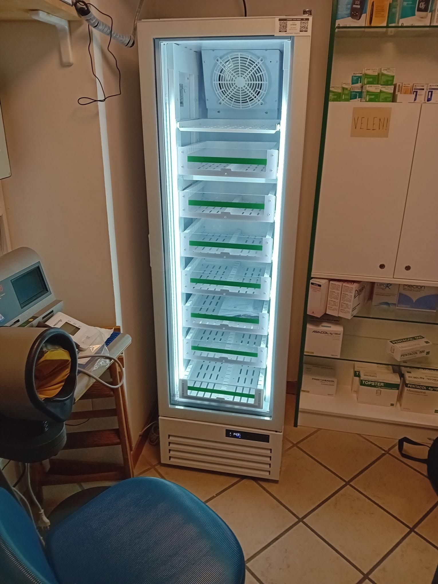 Frigorifero da laboratorio alto e illuminato, con vassoi impilati, accanto a una scrivania e armadietti in una stanza piastrellata.