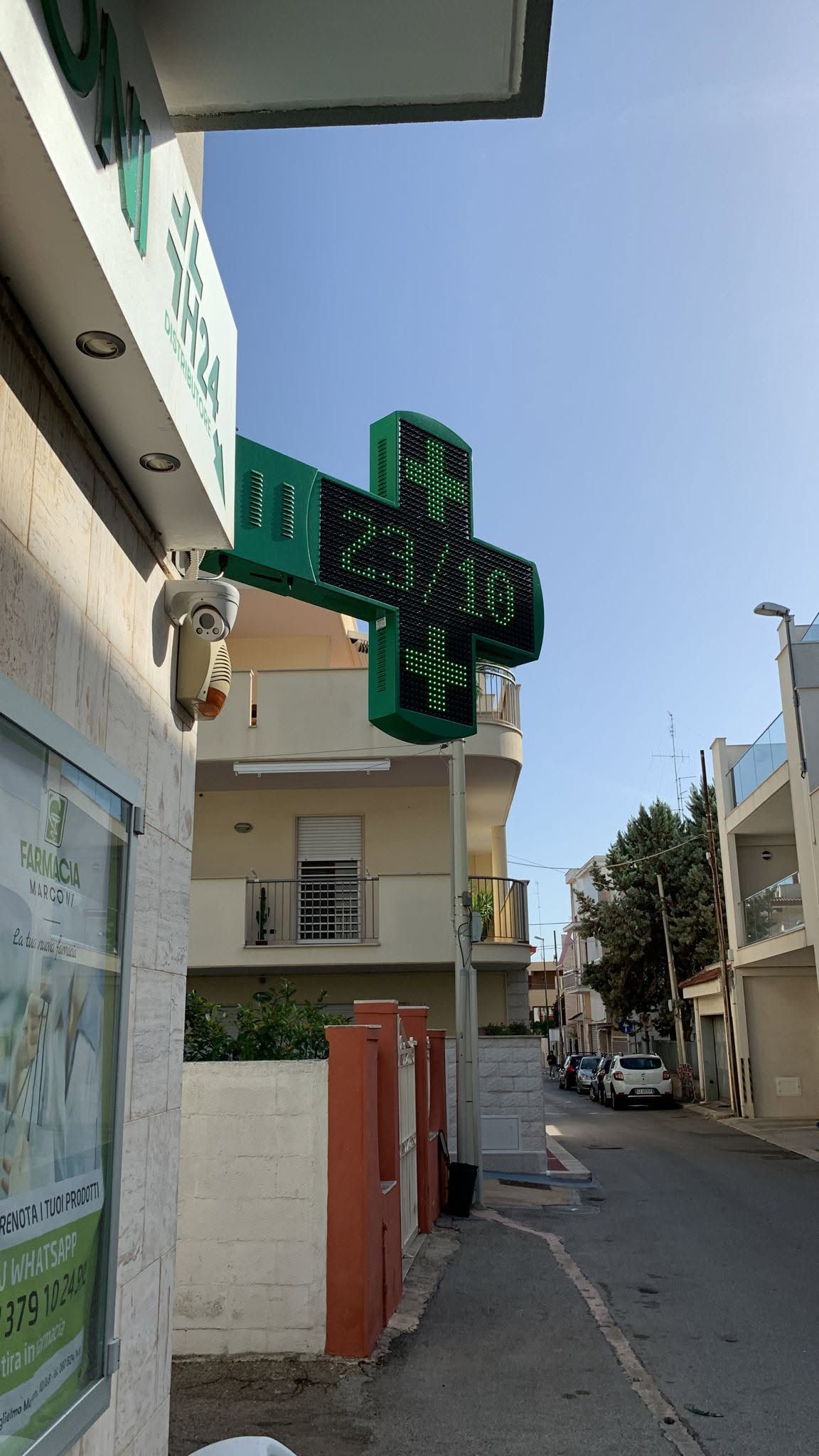 Stretta via fiancheggiata da edifici bassi, con un'insegna verde appesa e un cielo azzurro limpido.