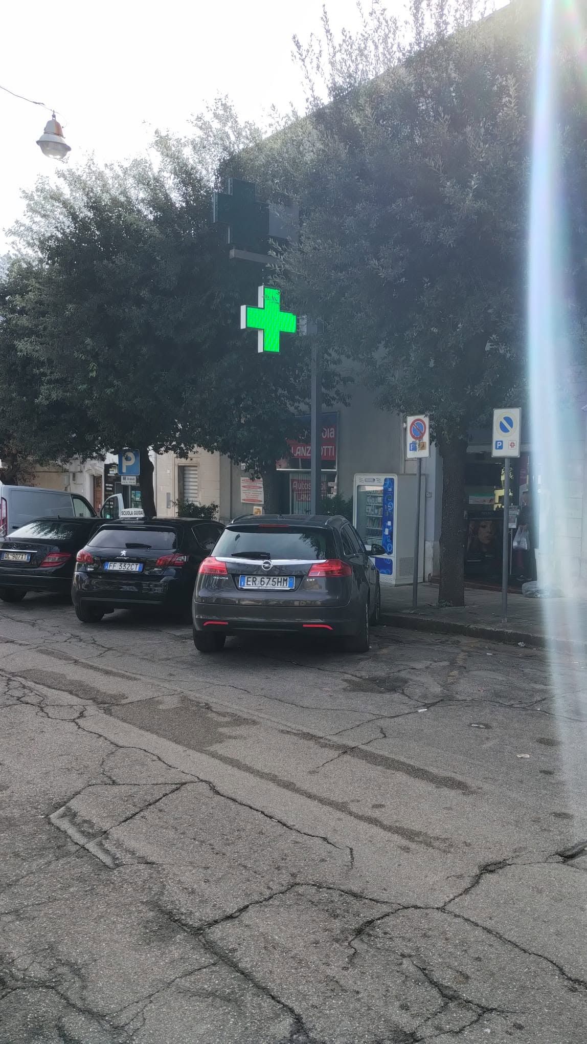 Scena di strada con auto parcheggiate, alberi e un'insegna a croce verde di una farmacia all'angolo.