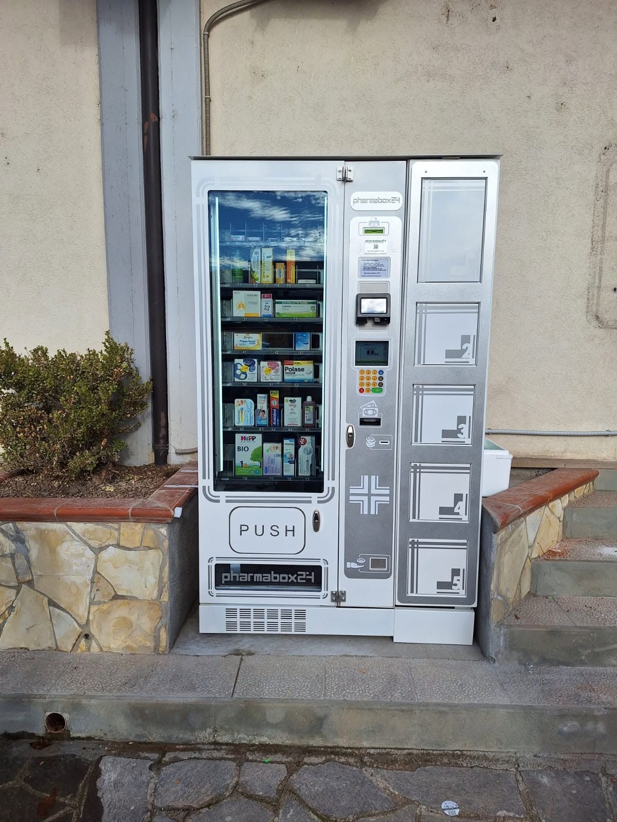 Distributore automatico bianco di bevande e snack situato all'esterno del muro di un edificio in pietra.