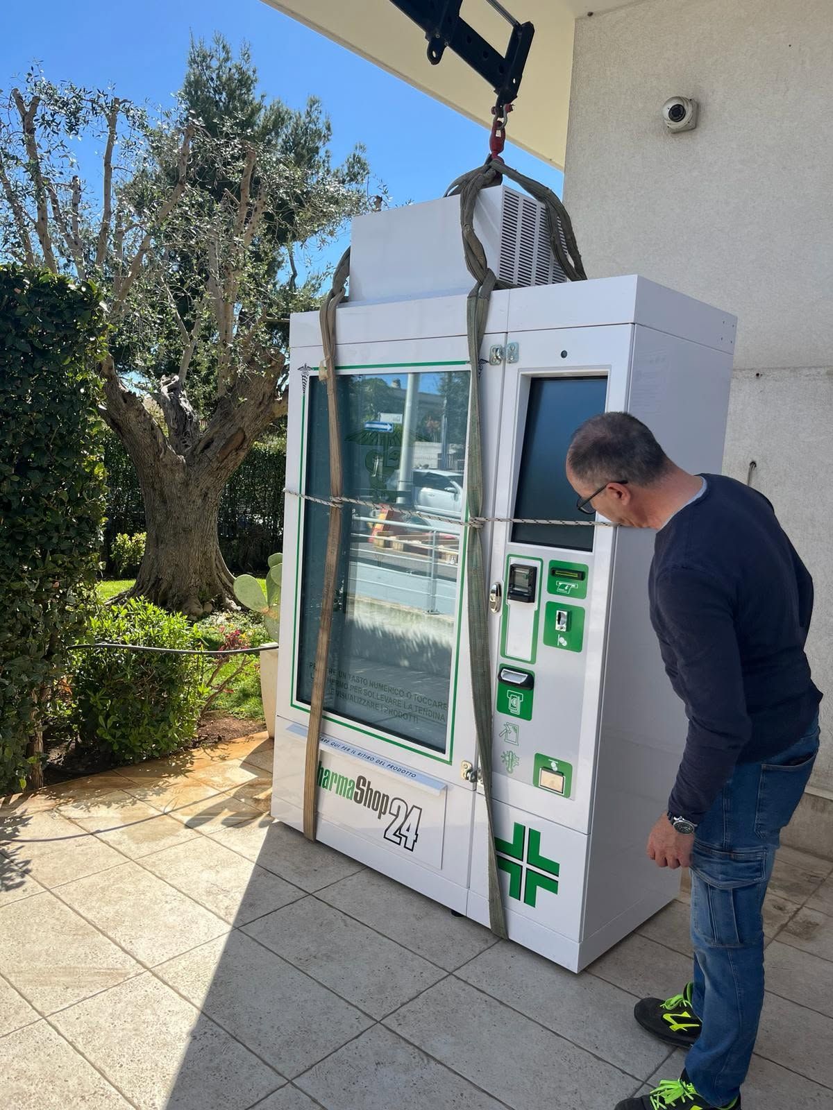 Un uomo esamina un distributore automatico bianco all'aperto, su un patio piastrellato, con un albero e il muro di un edificio nelle vicinanze.