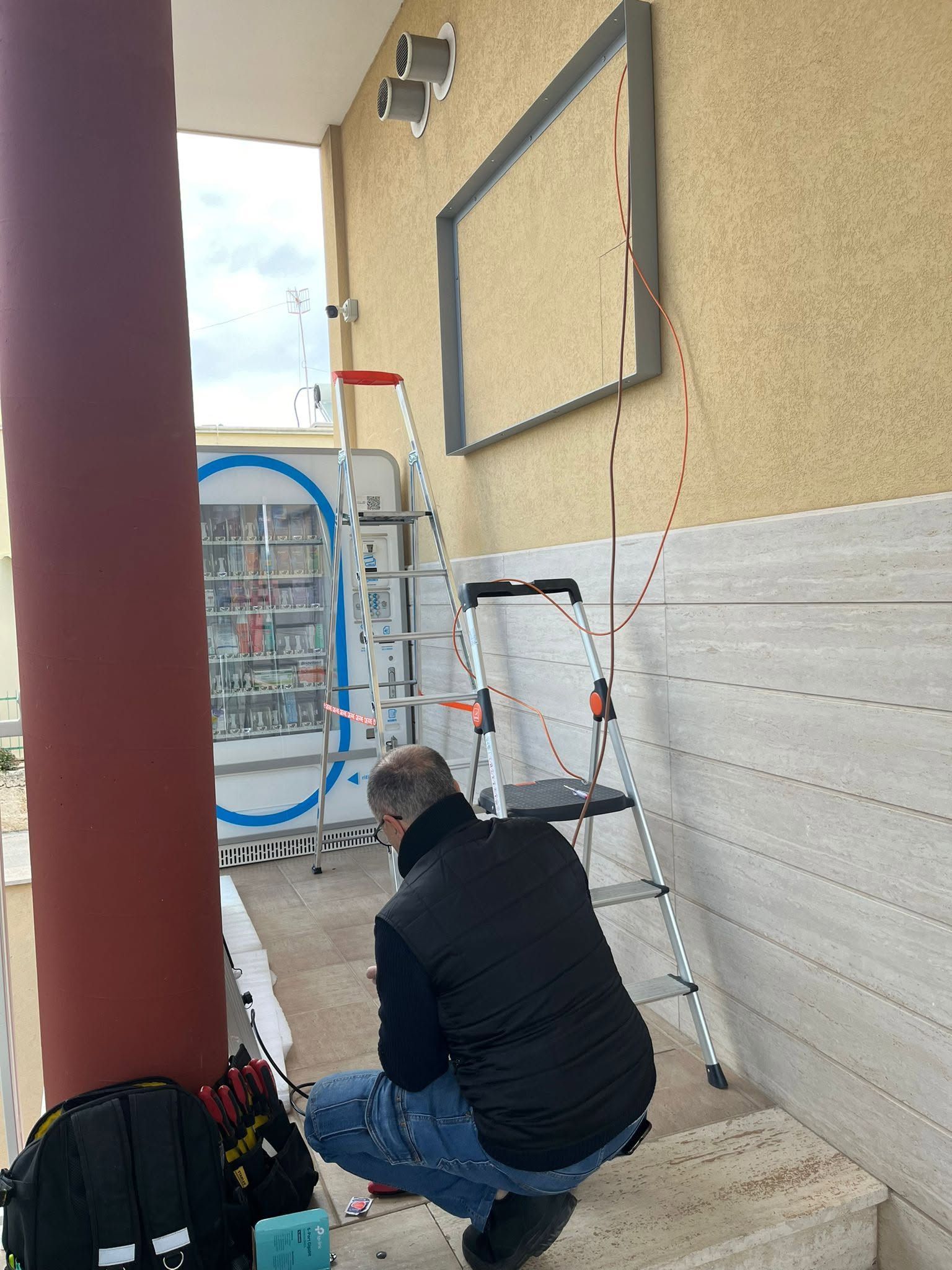 Una persona è inginocchiata accanto a una porta, intenta a lavorare con degli attrezzi vicino a una scala e a una colonna rossa all'ingresso di un edificio.