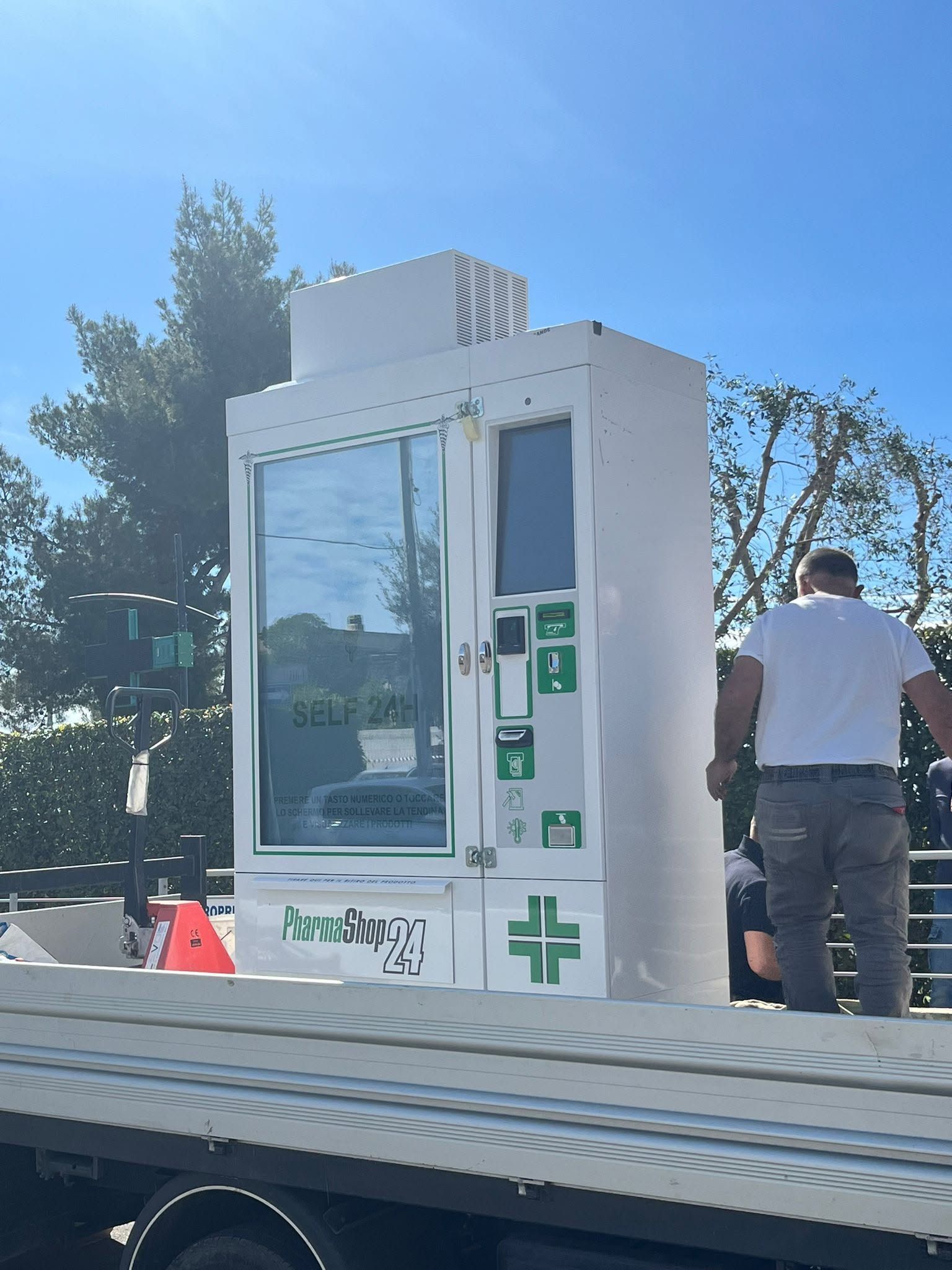 Chiosco bianco a bordo strada con simboli a croce verdi; un uomo è in piedi accanto ad esso all'aperto sotto un cielo azzurro.