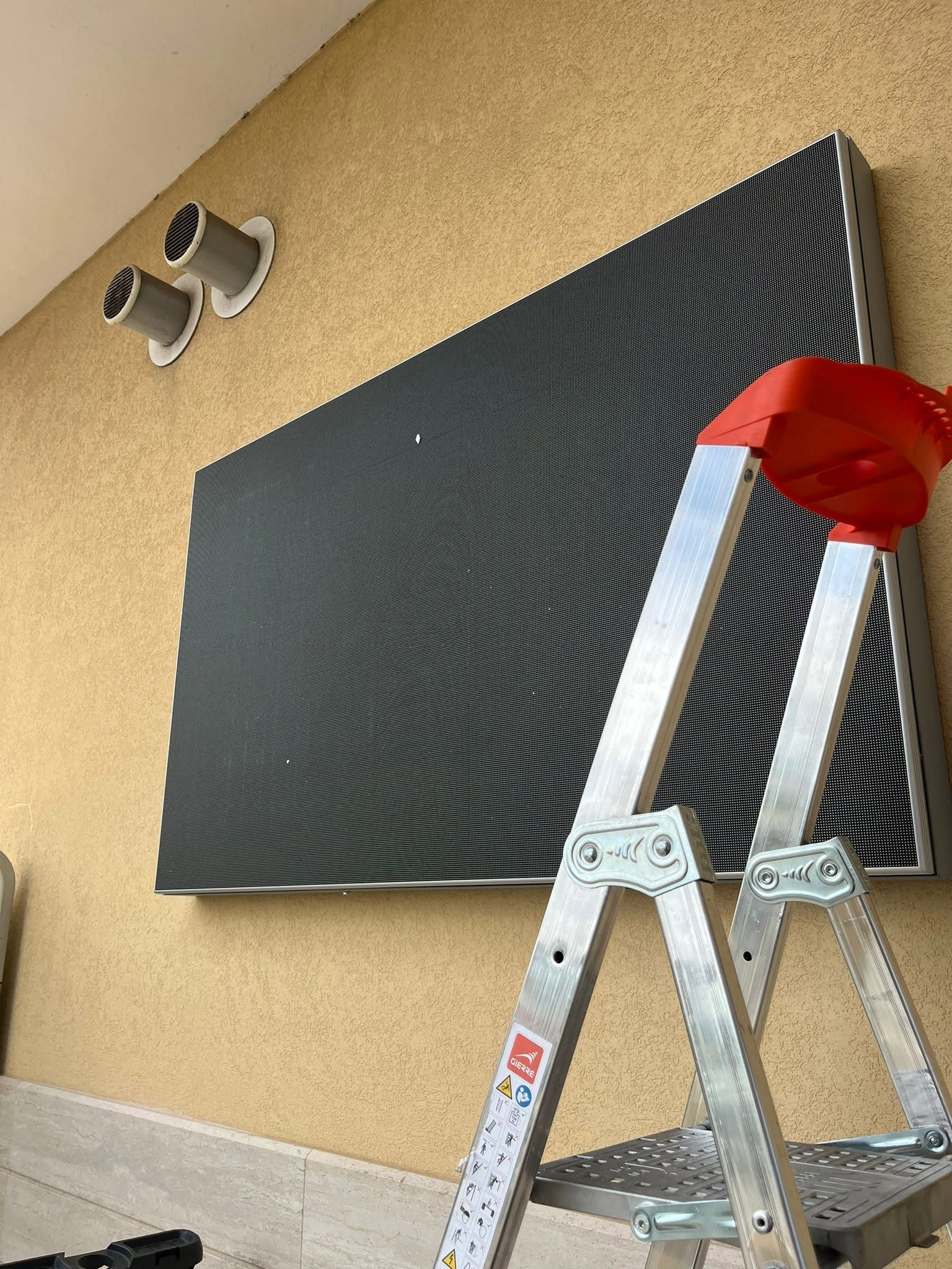 Scala accanto a un pannello acustico nero montato su una parete esterna color beige, vicino a una presa d'aria.