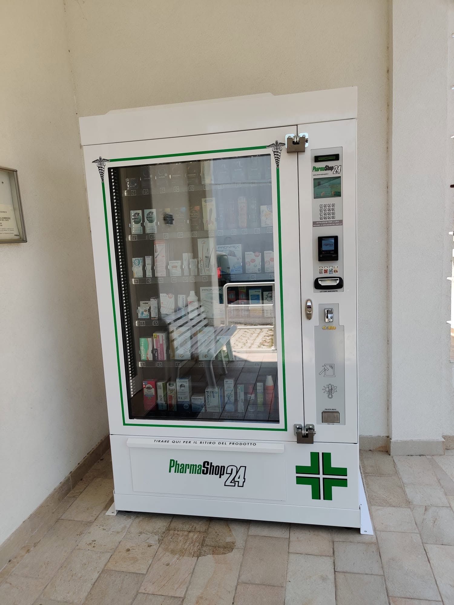 Distributore automatico bianco in un corridoio, con frontale in vetro, tastiera e logo a croce verde.