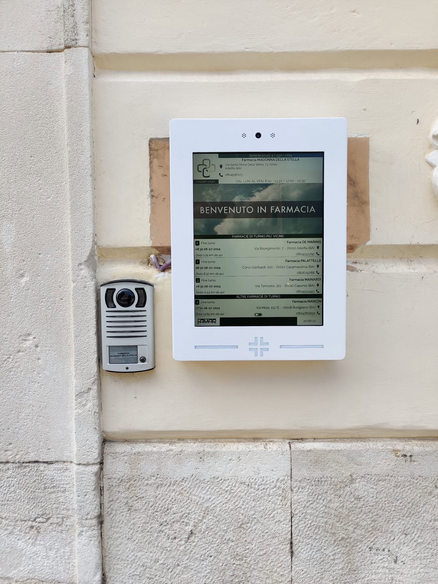 Schermo digitale per le informazioni e citofono montati a parete sulla facciata di un edificio in pietra beige.