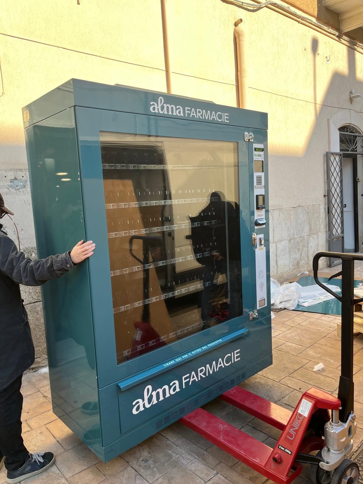 Un distributore automatico blu di farmaci viene scaricato all'aperto da un operaio che utilizza un transpallet rosso.