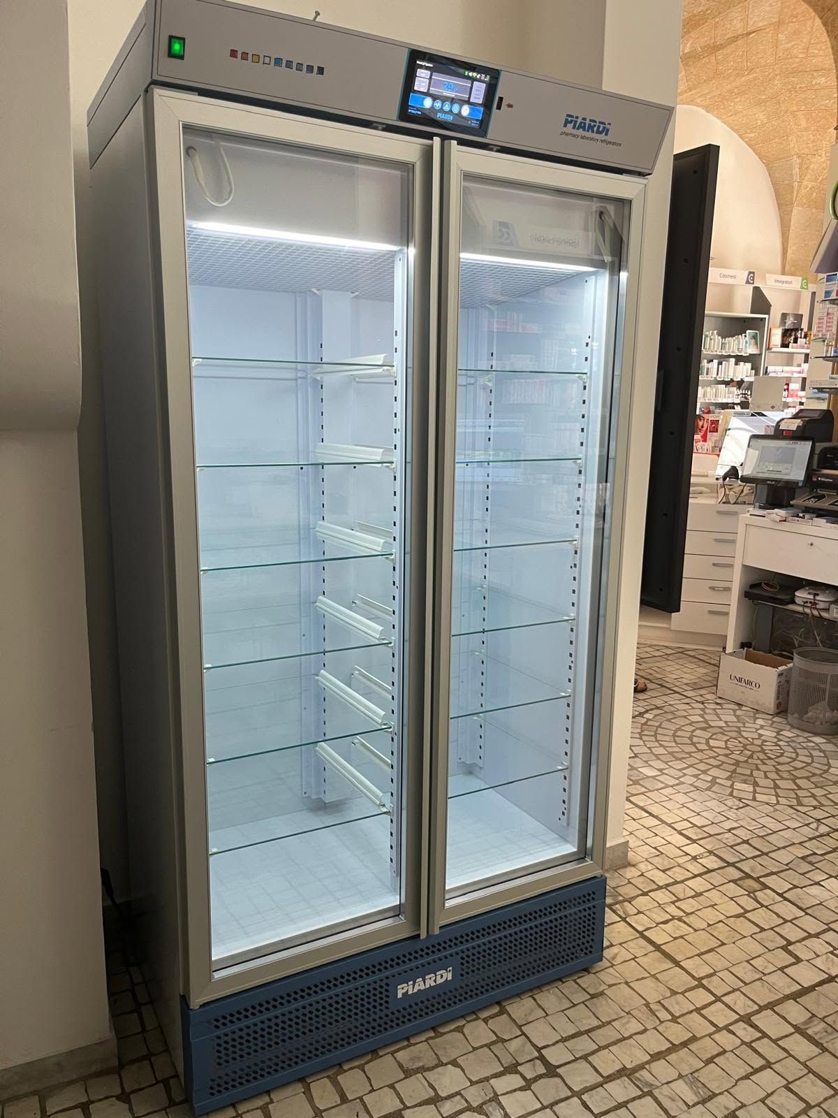 Frigorifero commerciale vuoto con porta in vetro in una stanza piastrellata con ripiani interni bianchi e base blu