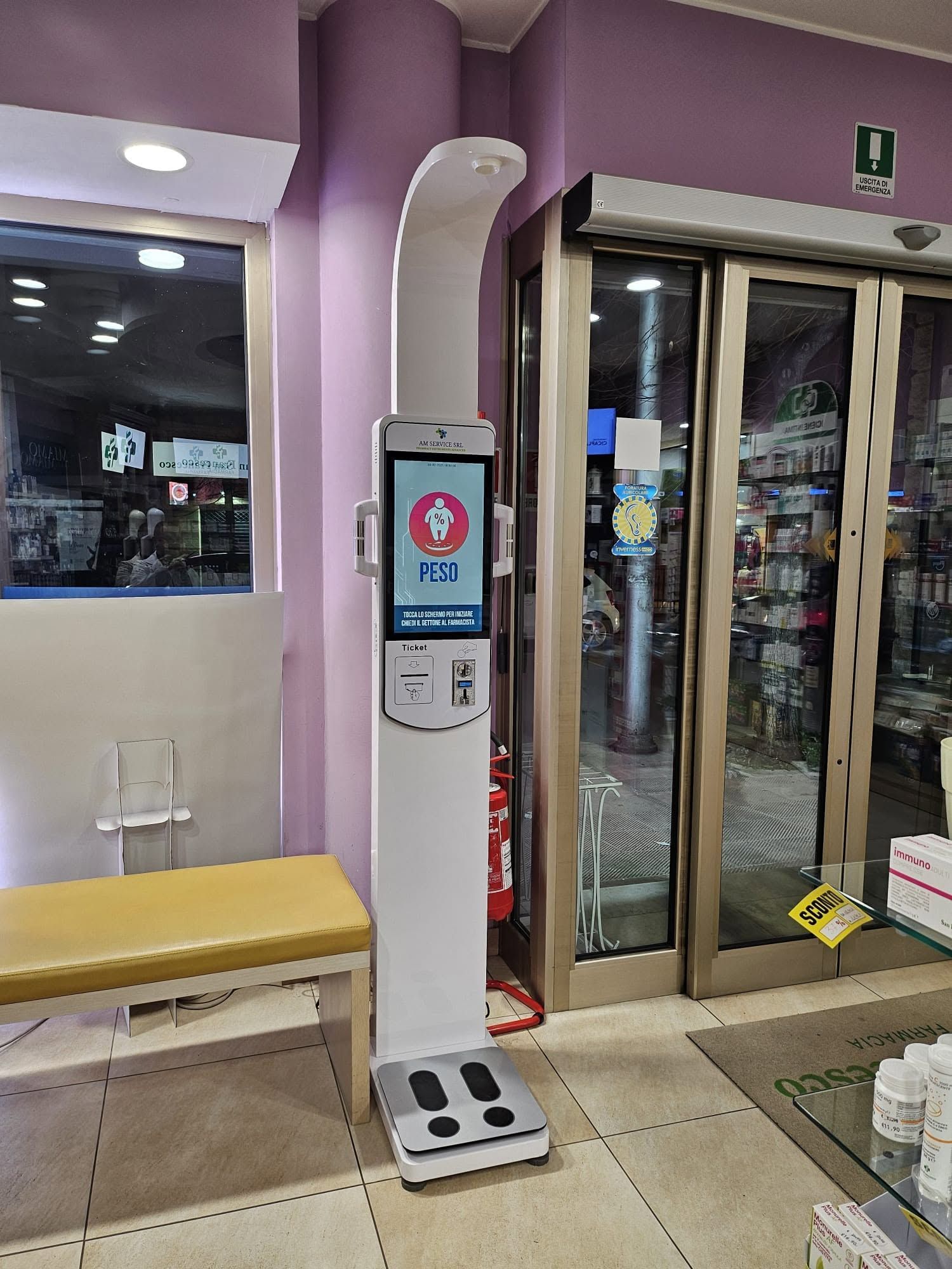 Ingresso interno con porte a vetri automatiche, postazione per la disinfezione delle mani, panchina e parete viola.