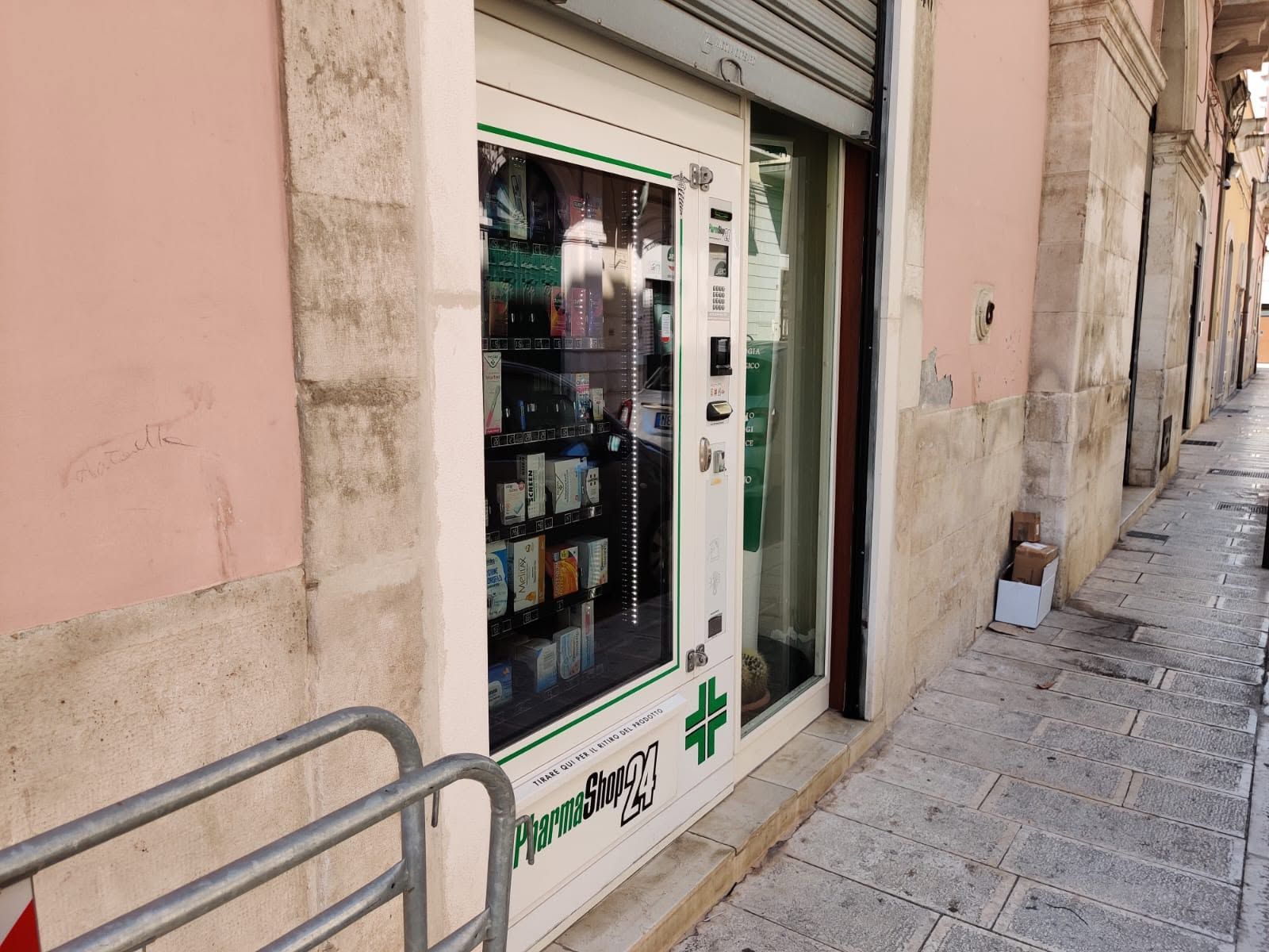 Piccola farmacia con insegna a croce verde, situata su una stretta via lastricata.