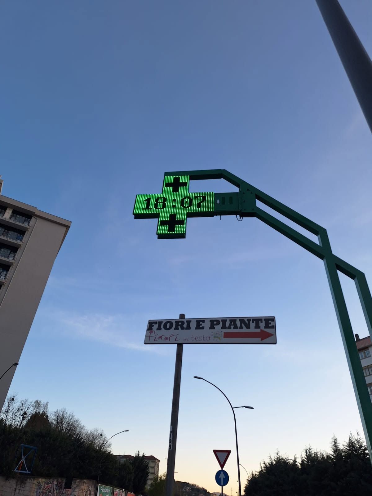 Una croce digitale verde su un palo sopra un segnale stradale, con cielo azzurro ed edifici al crepuscolo.