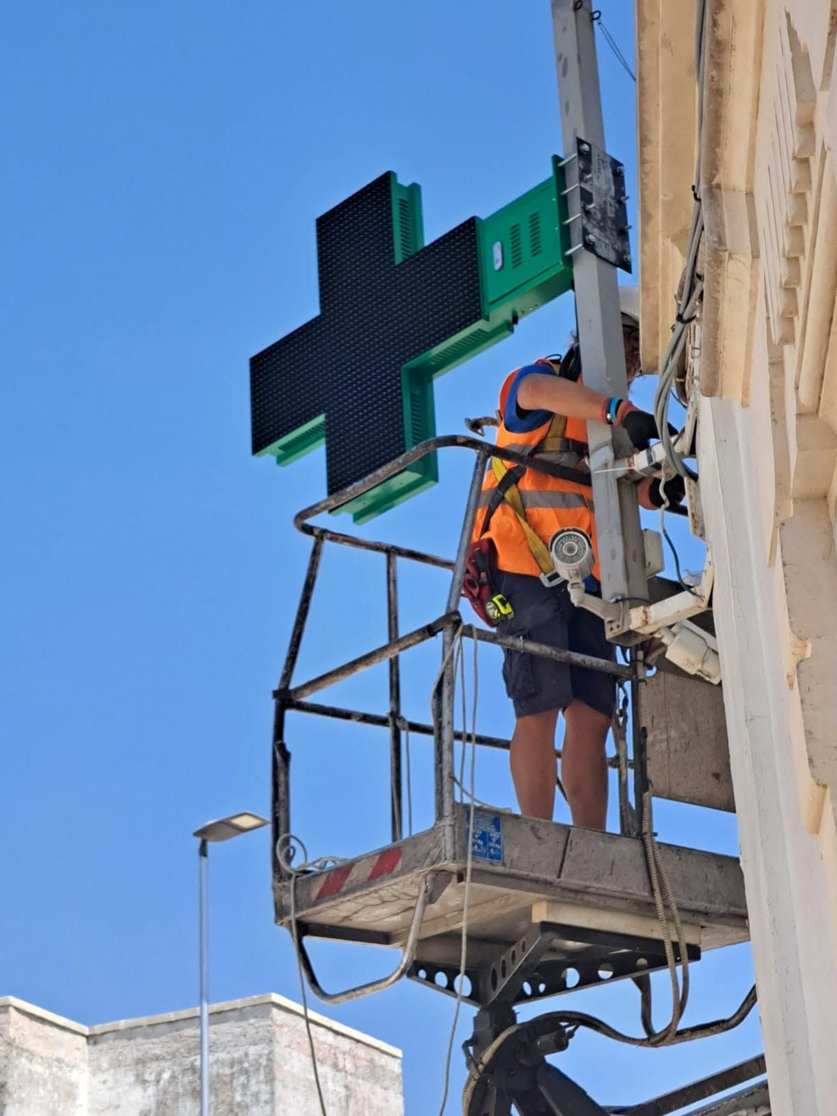 Un operaio con giubbotto di sicurezza arancione su una piattaforma elevatrice sta riparando un alto semaforo sullo sfondo di un cielo azzurro.