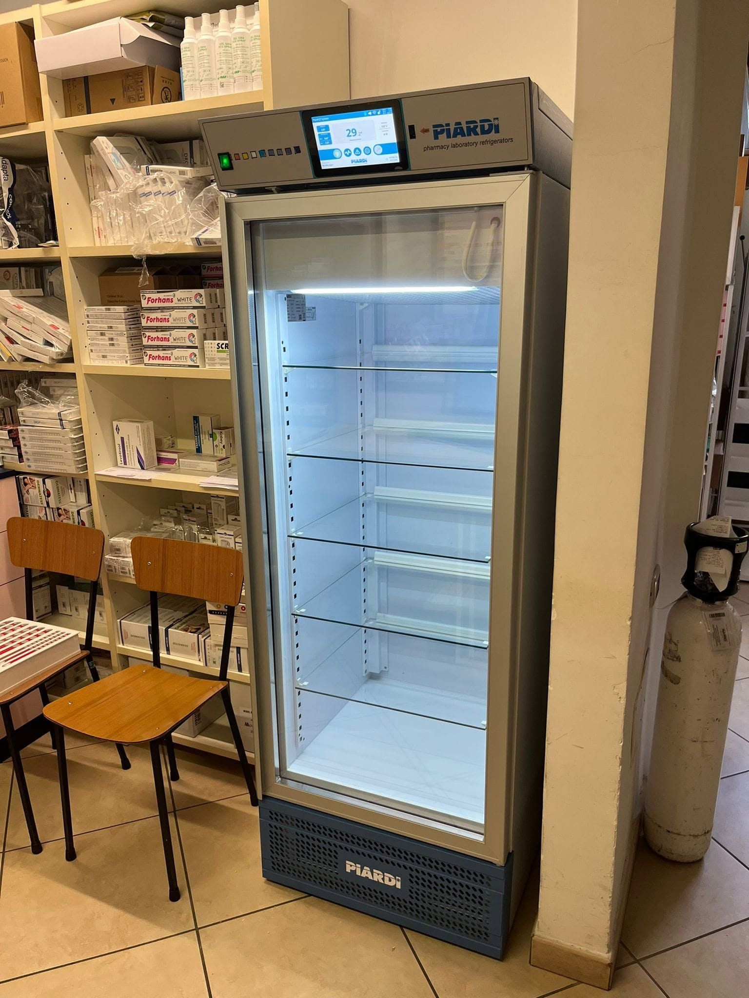 Frigorifero vuoto con porta in vetro in un piccolo negozio o bar, con scaffali e sedie accanto.