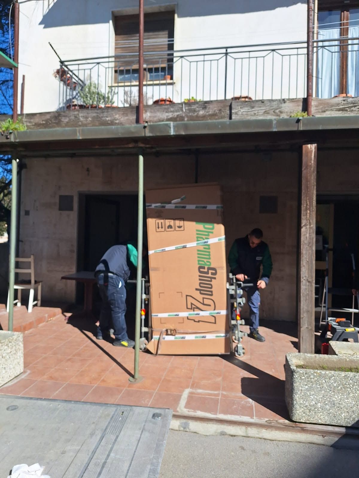 Due persone spostano una grande scatola di cartone attraverso una porta con telaio metallico verde su un patio di mattoni.