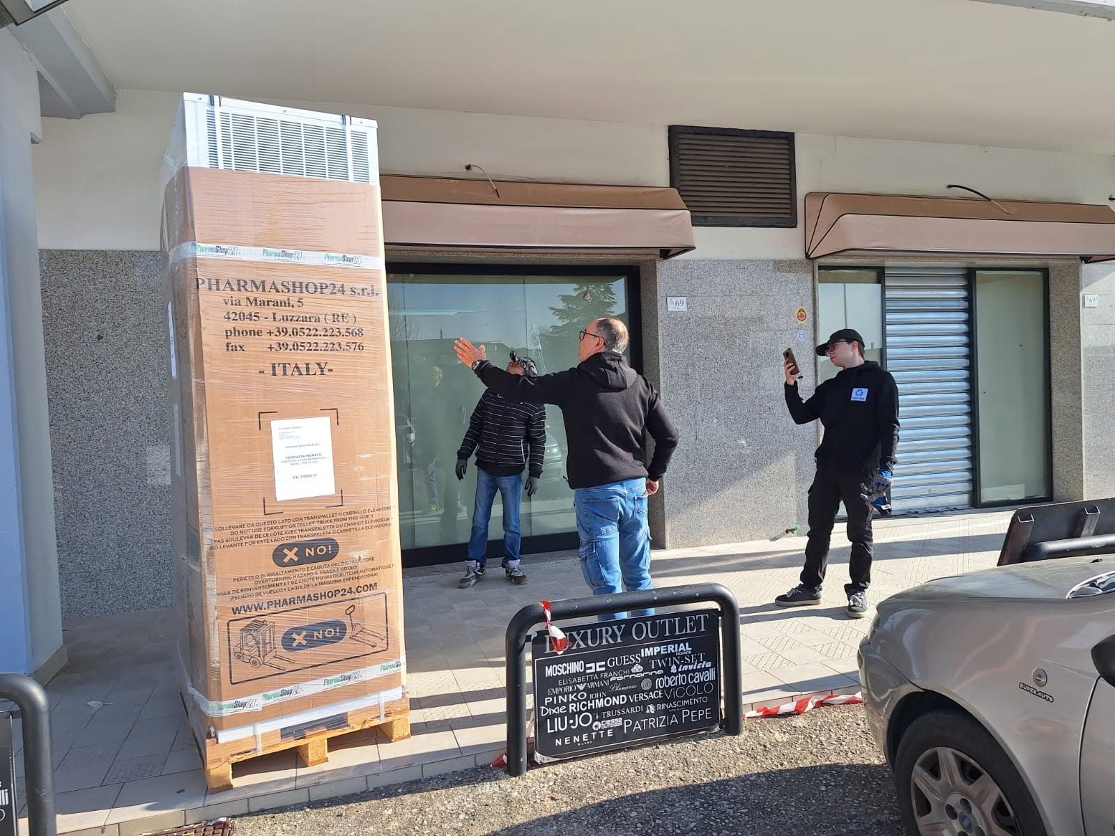 Tre persone osservano un murale accanto a un grande pallet avvolto in un involucro, sul marciapiede di un negozio.