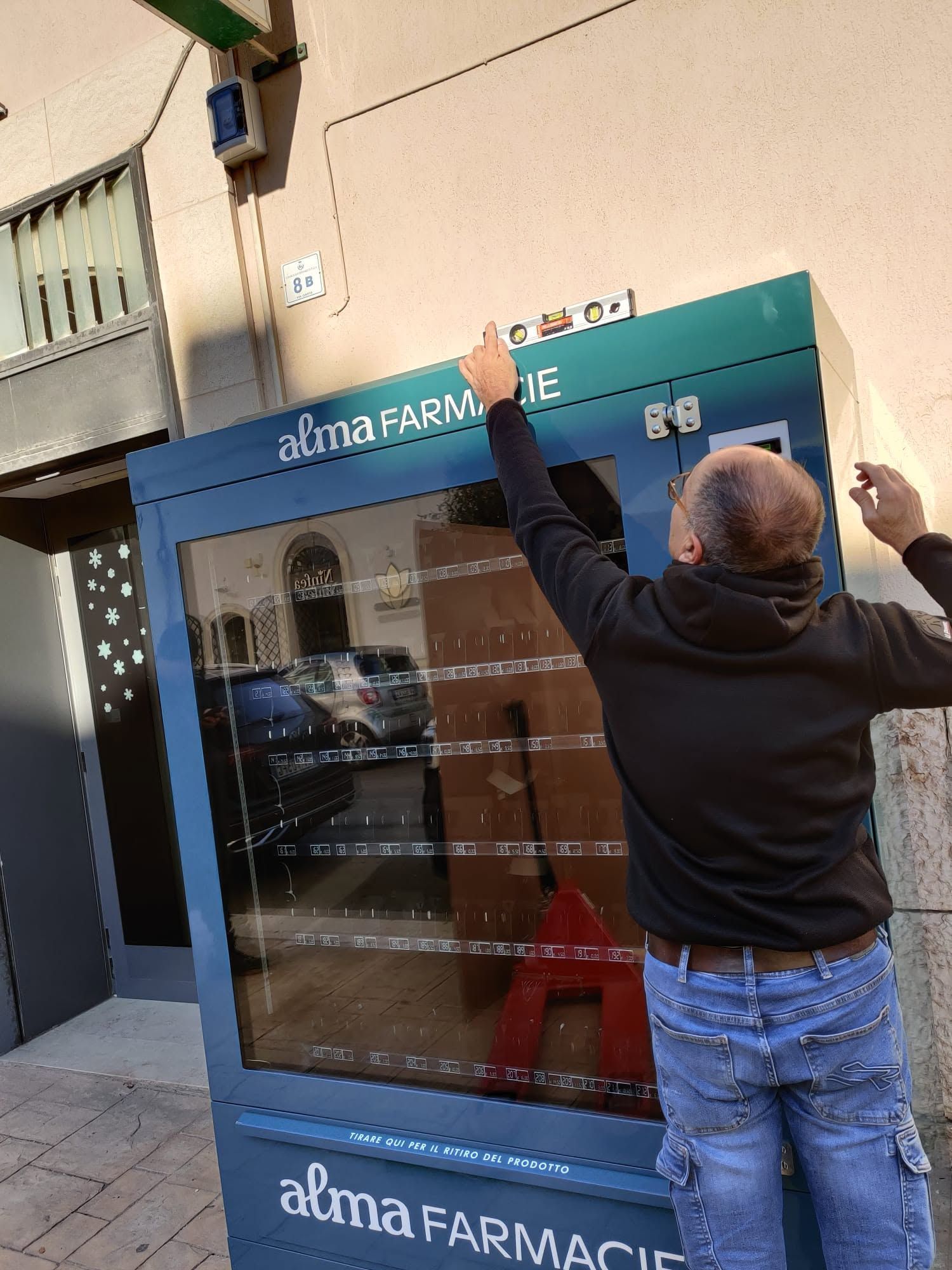 Una persona si allunga per sistemare l'insegna sulla vetrina blu di un negozio Alma Farmacia.