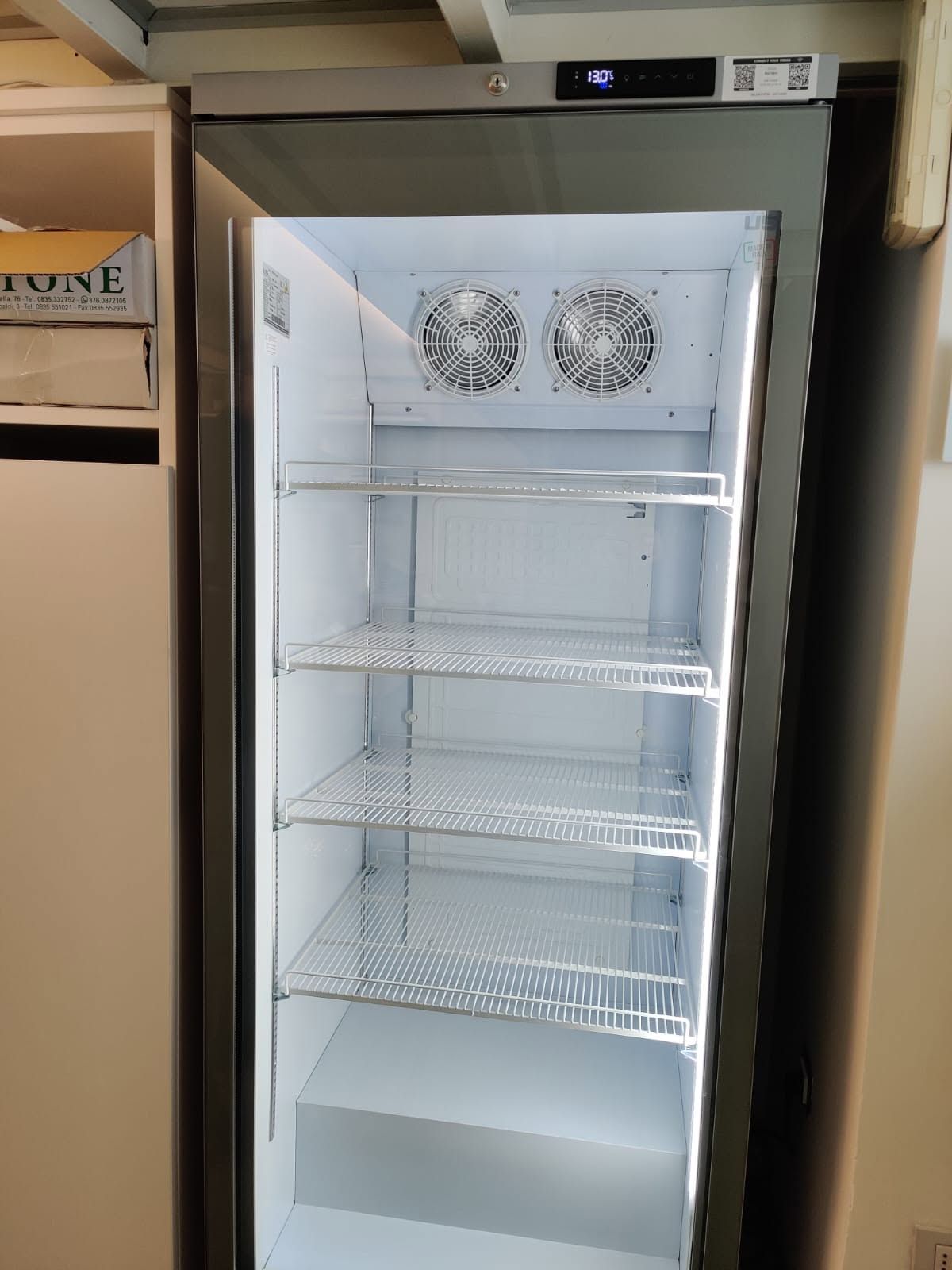 Frigorifero commerciale aperto con ripiani e due ventole all'interno, con la porta aperta in cucina o in negozio.