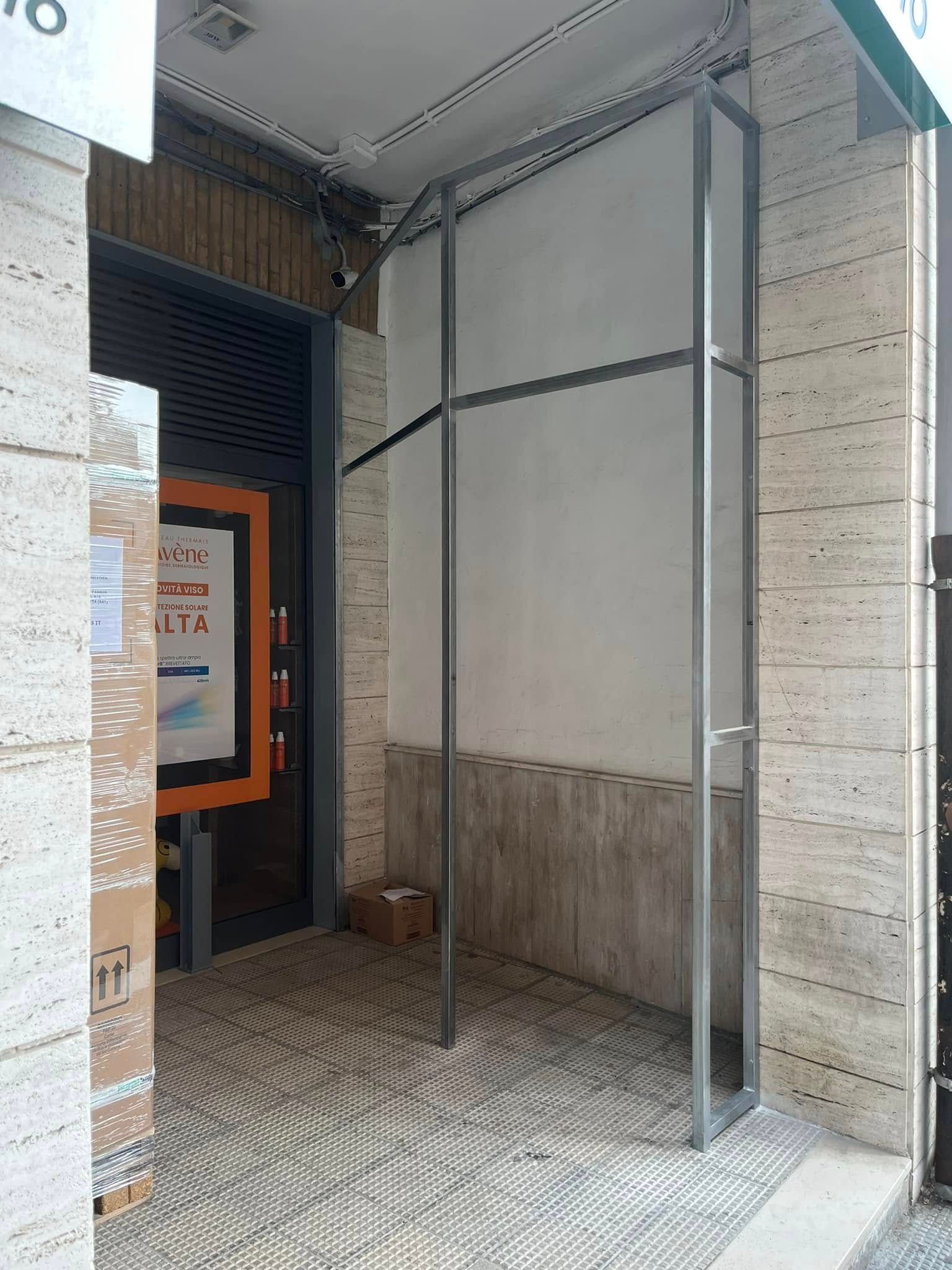 Ingresso coperto dell'edificio con struttura metallica, pavimento piastrellato e un poster sulla parete sinistra.