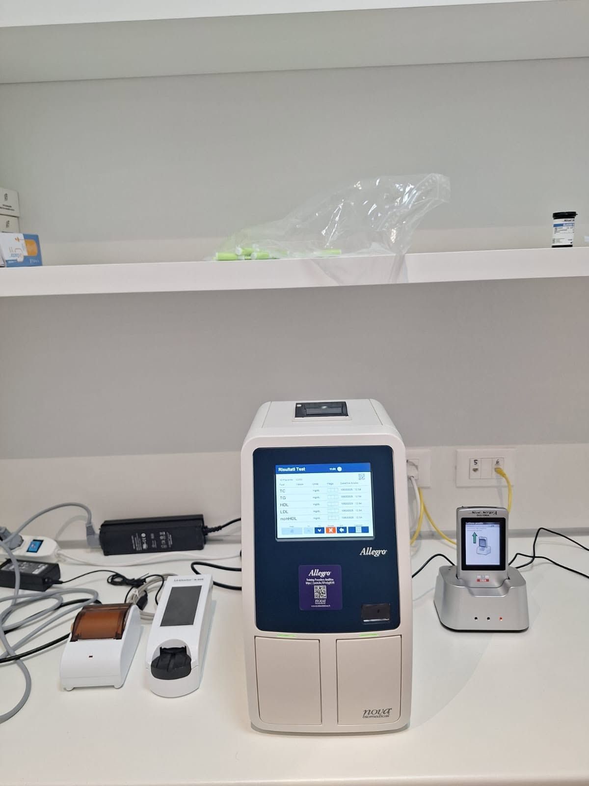Banco da laboratorio con un analizzatore medico bianco, uno smartphone e dei cavi, appoggiati a una parete spoglia.