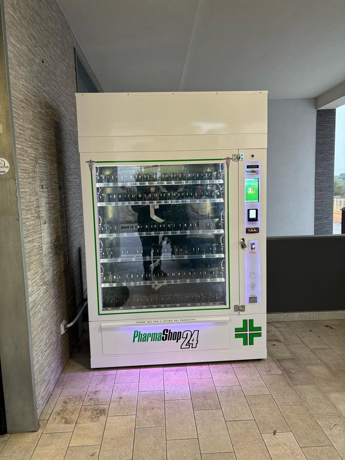 Distributore automatico beige in un corridoio, con ripiani illuminati e logo a croce verde.