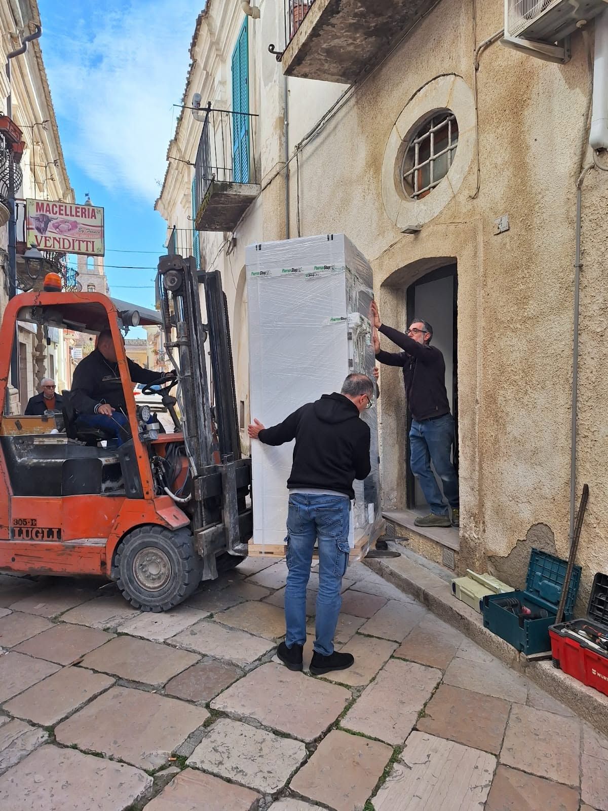 Operai che caricano un grosso oggetto bianco su un carrello elevatore in uno stretto vicolo lastricato in pietra.