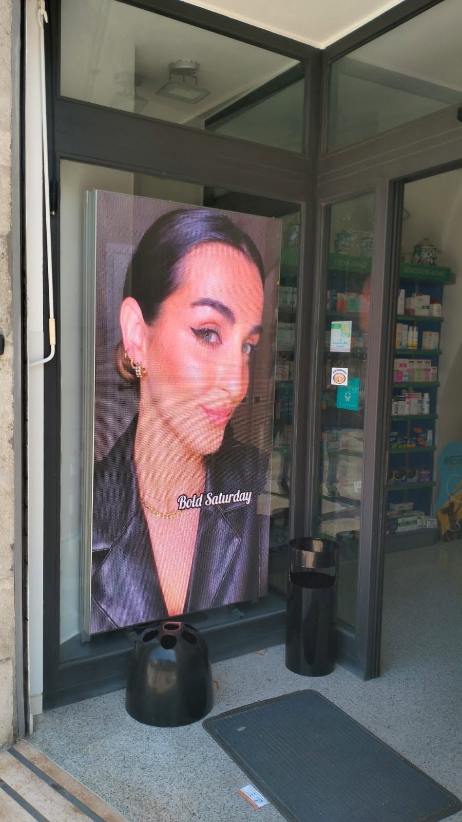 Un grande poster raffigurante il volto di una donna sull'ingresso di un negozio con porte a vetri e uno zerbino nero.