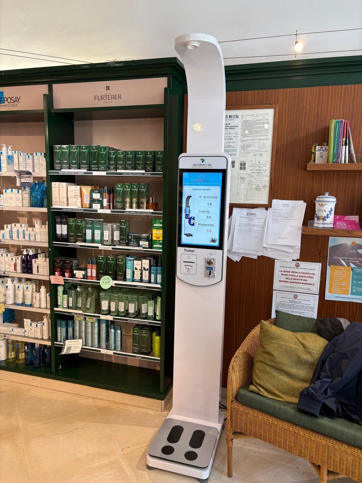 Un alto chiosco touchscreen bianco si trova in una corsia di un negozio, accanto a scaffali di prodotti e a una sedia in vimini.