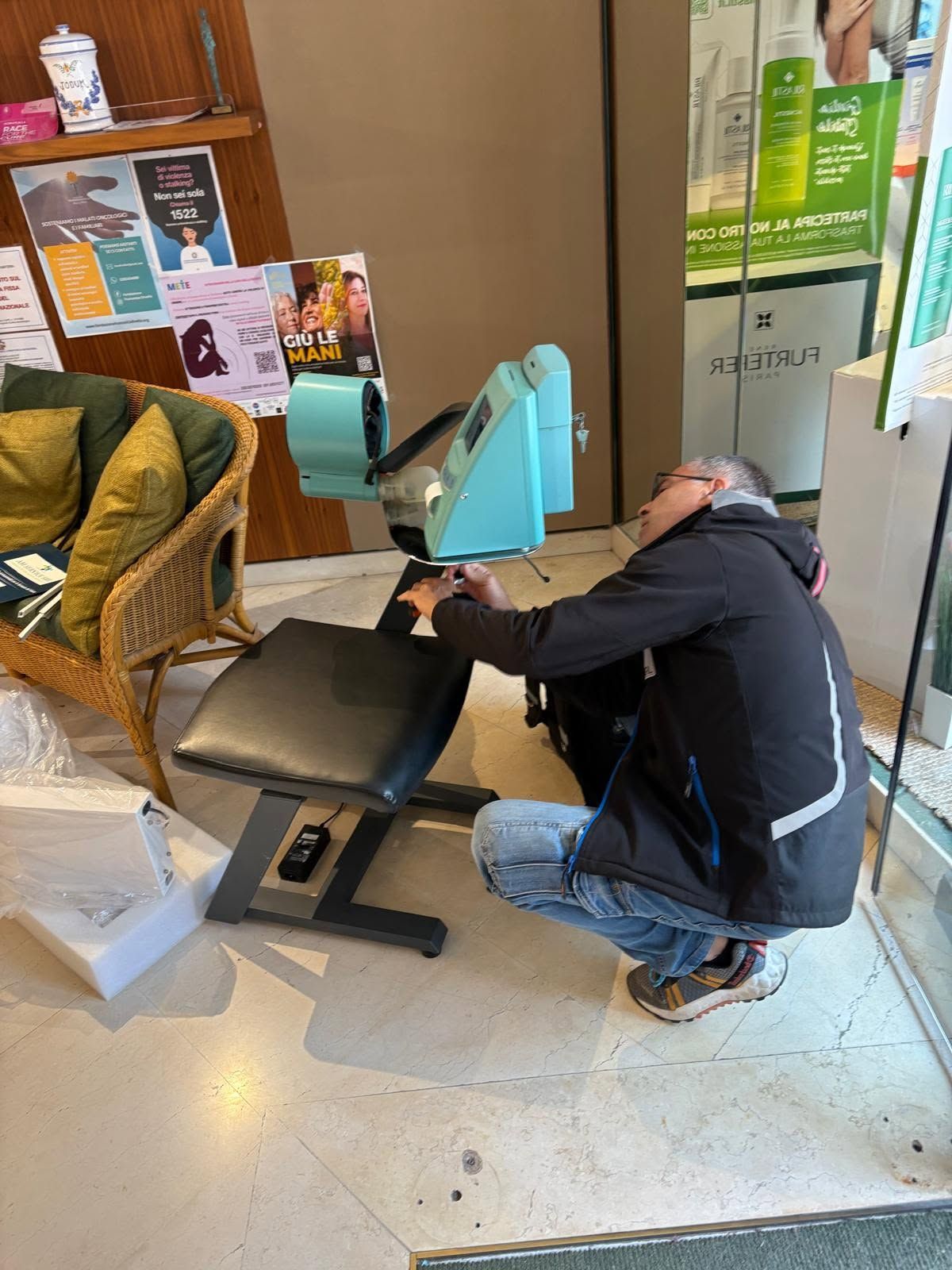 Una persona inginocchiata accanto a una sedia nera, intenta a regolare una macchina color verde acqua in uno showroom o nella hall di un ufficio.