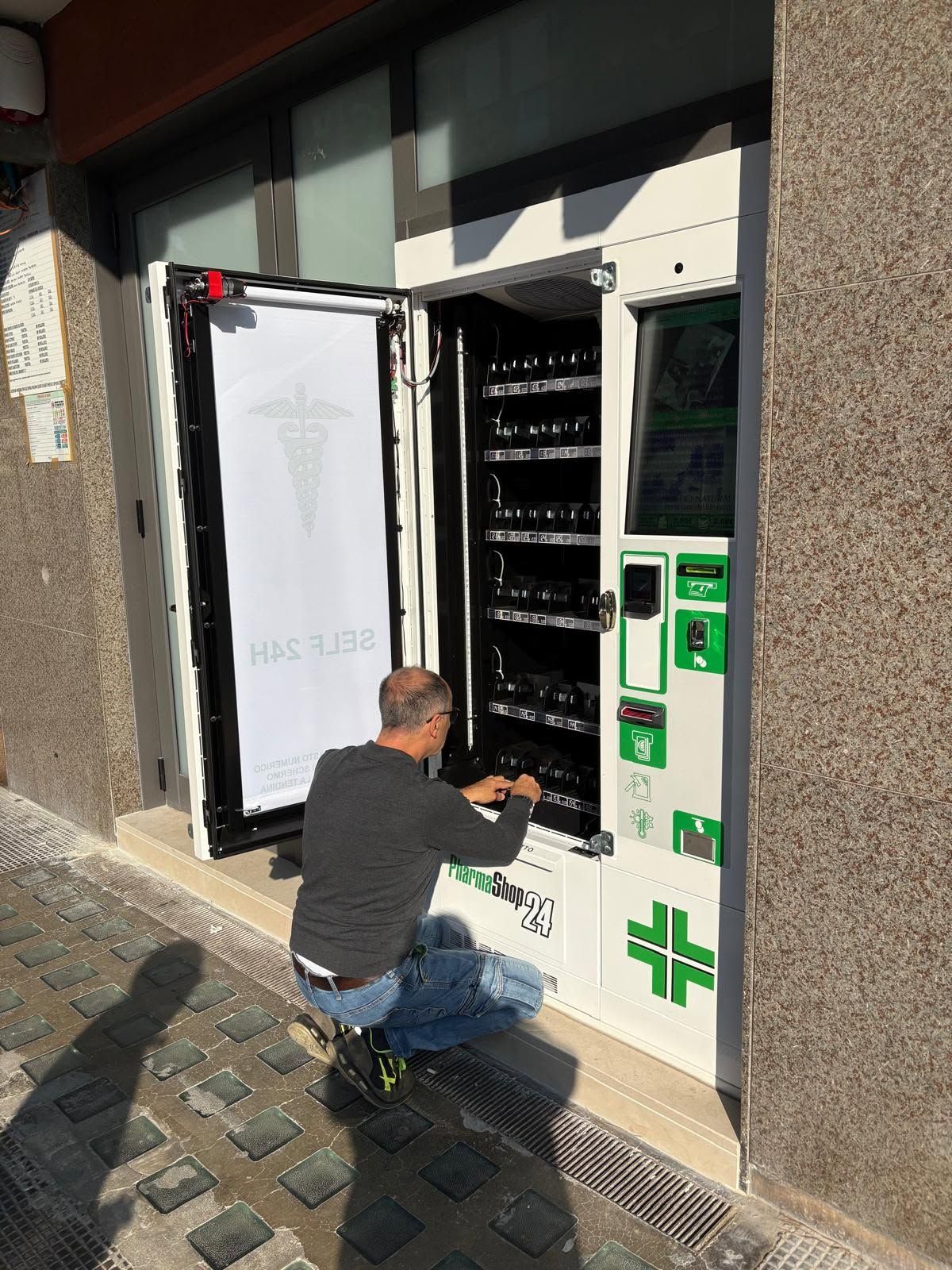 Un uomo è inginocchiato accanto a un distributore automatico all'aperto, intento a sistemare gli scaffali vicino a una vetrina.