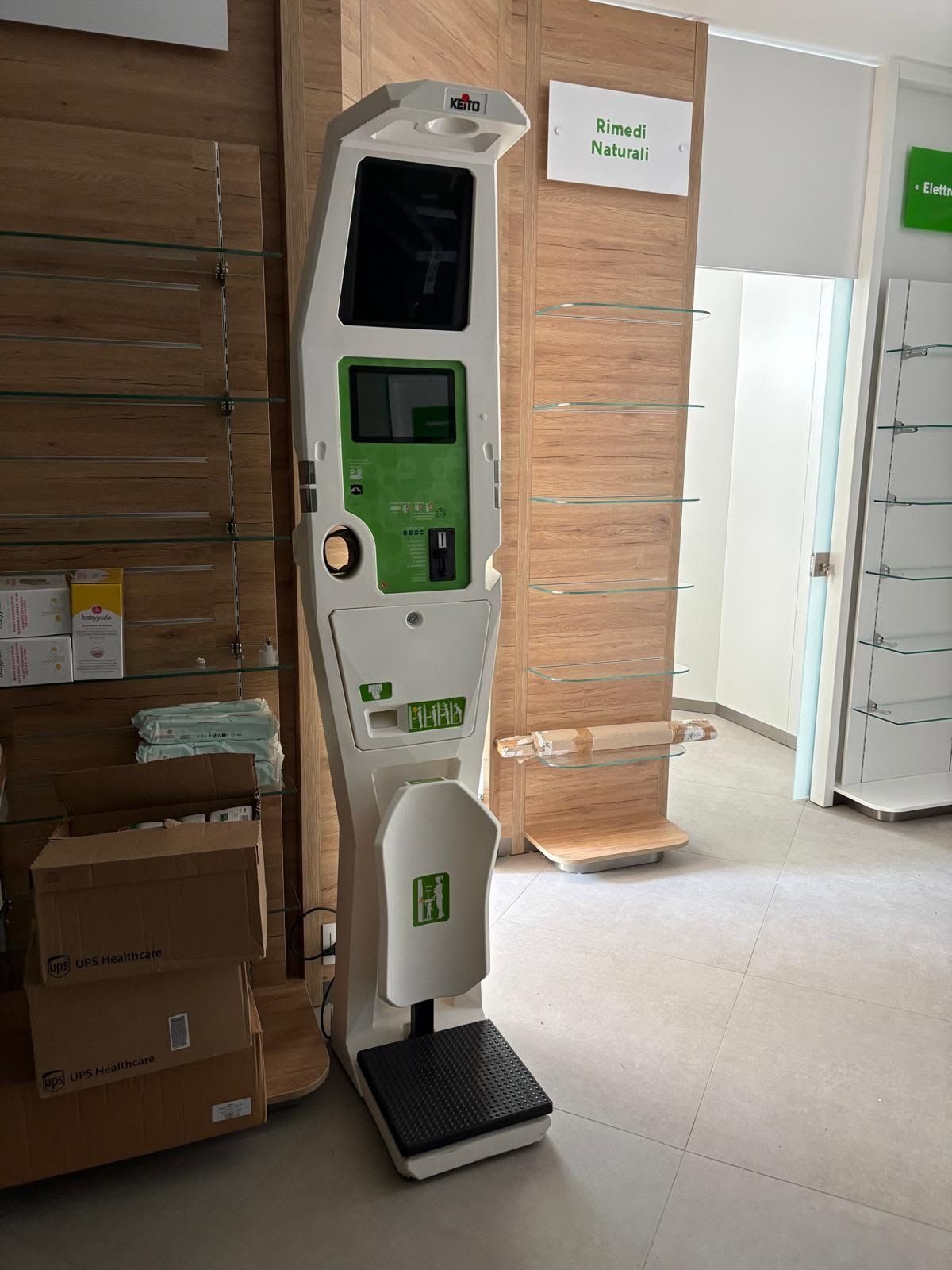 Chiosco self-service con schermo e tastiera in una corsia di un negozio, accanto a scatole di cartone e assi impilate.
